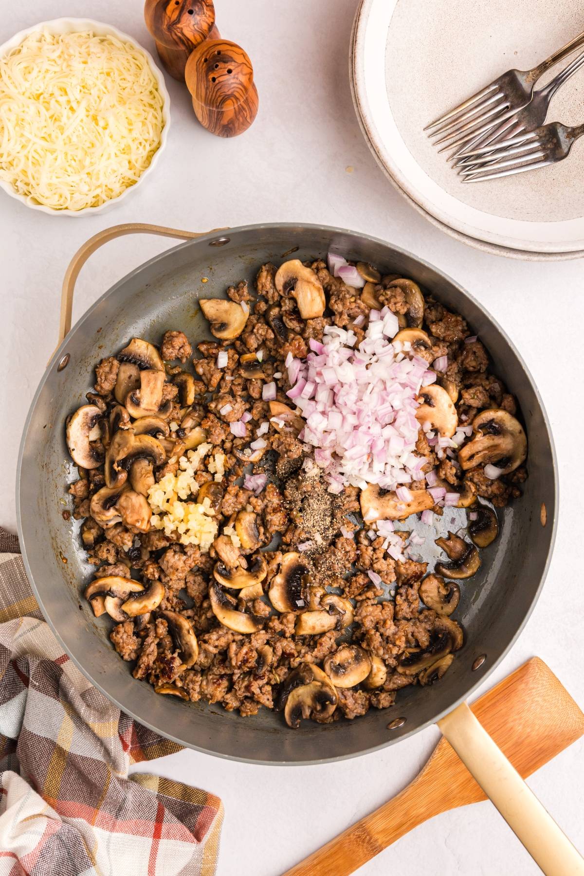 A skillet with cooked ground beef, mushrooms, chopped onions, and garlic, with cheese and plates nearby.