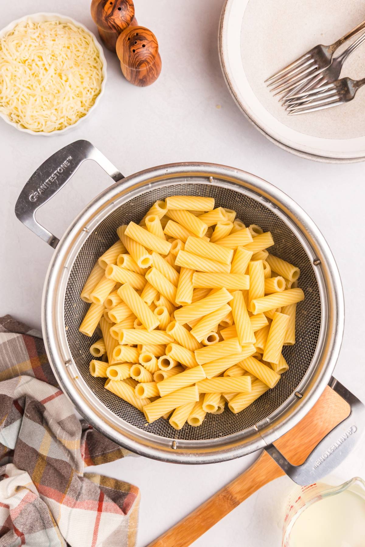 Cooked rigatoni pasta in a metal strainer, with cheese, plates, and forks nearby on a light countertop.