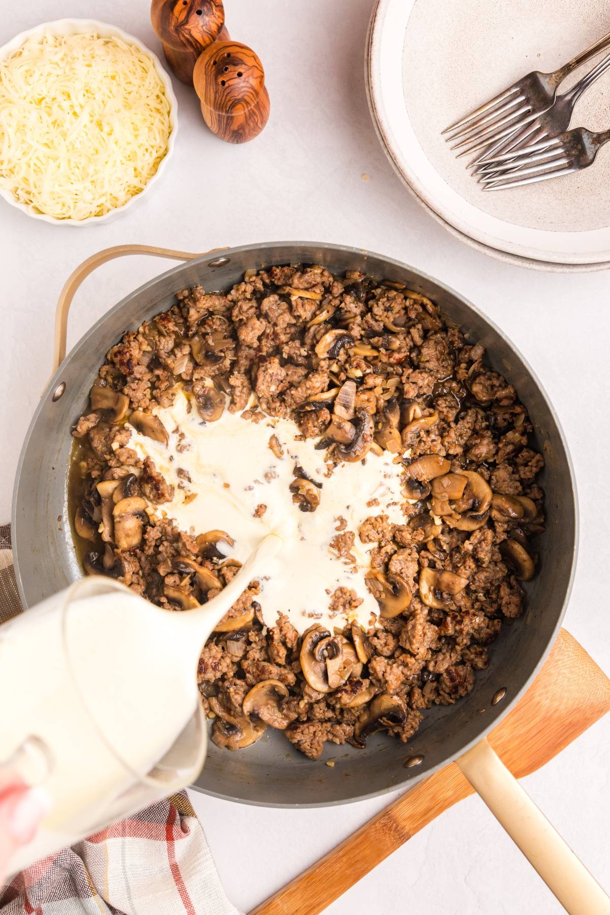 Cream is being poured into a skillet with cooked ground beef and mushrooms; cheese and plates are nearby.
