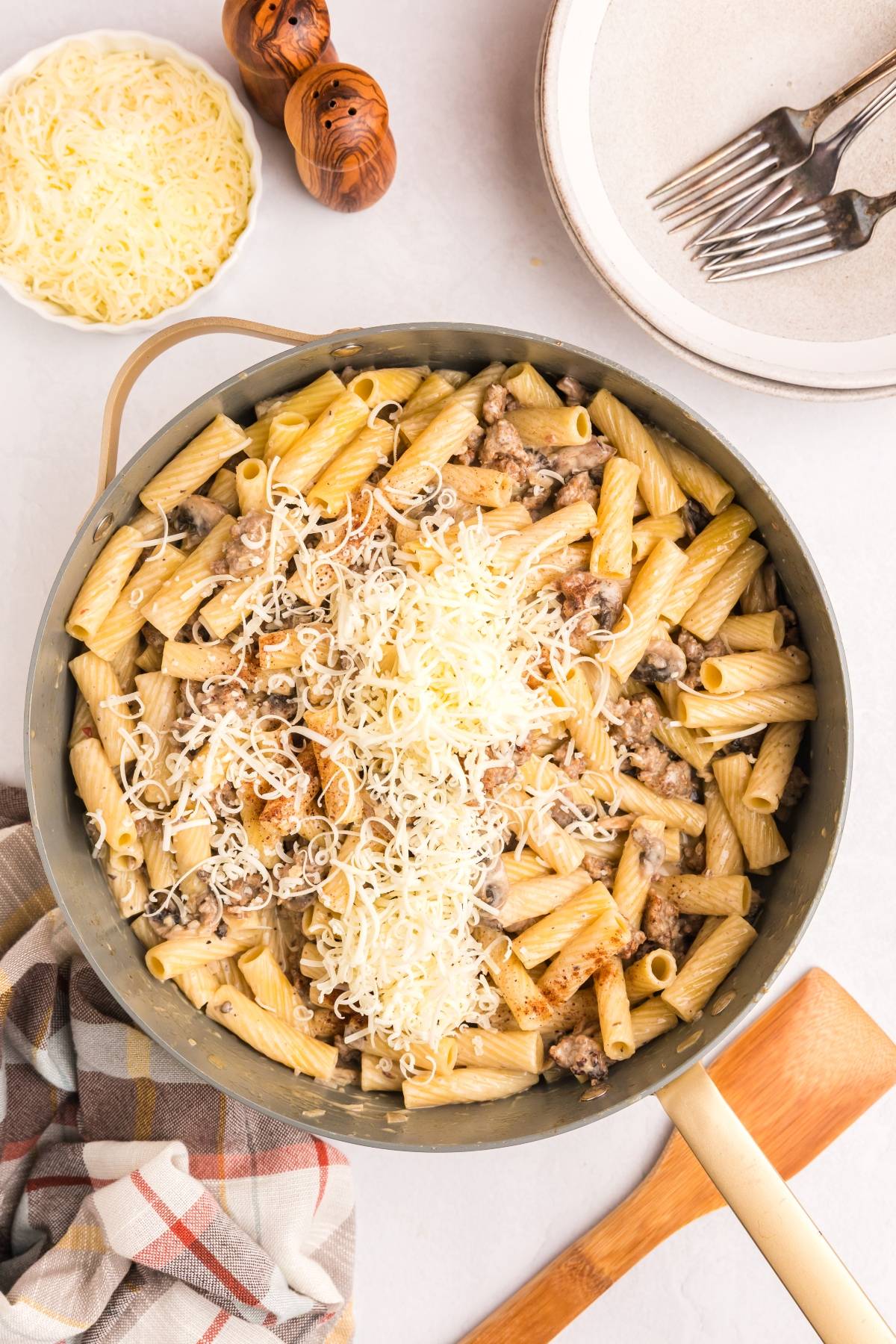 A skillet of rigatoni pasta with sausage and shredded cheese, surrounded by plates, forks, and a bowl of cheese.