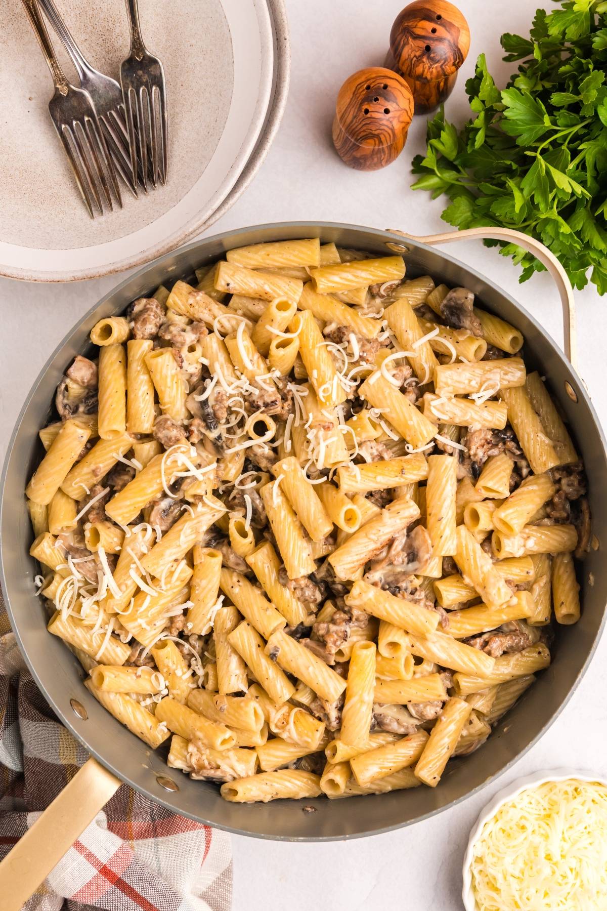 A skillet of creamy rigatoni pasta with mushrooms and cheese, next to plates, forks, parsley, and cheese.
