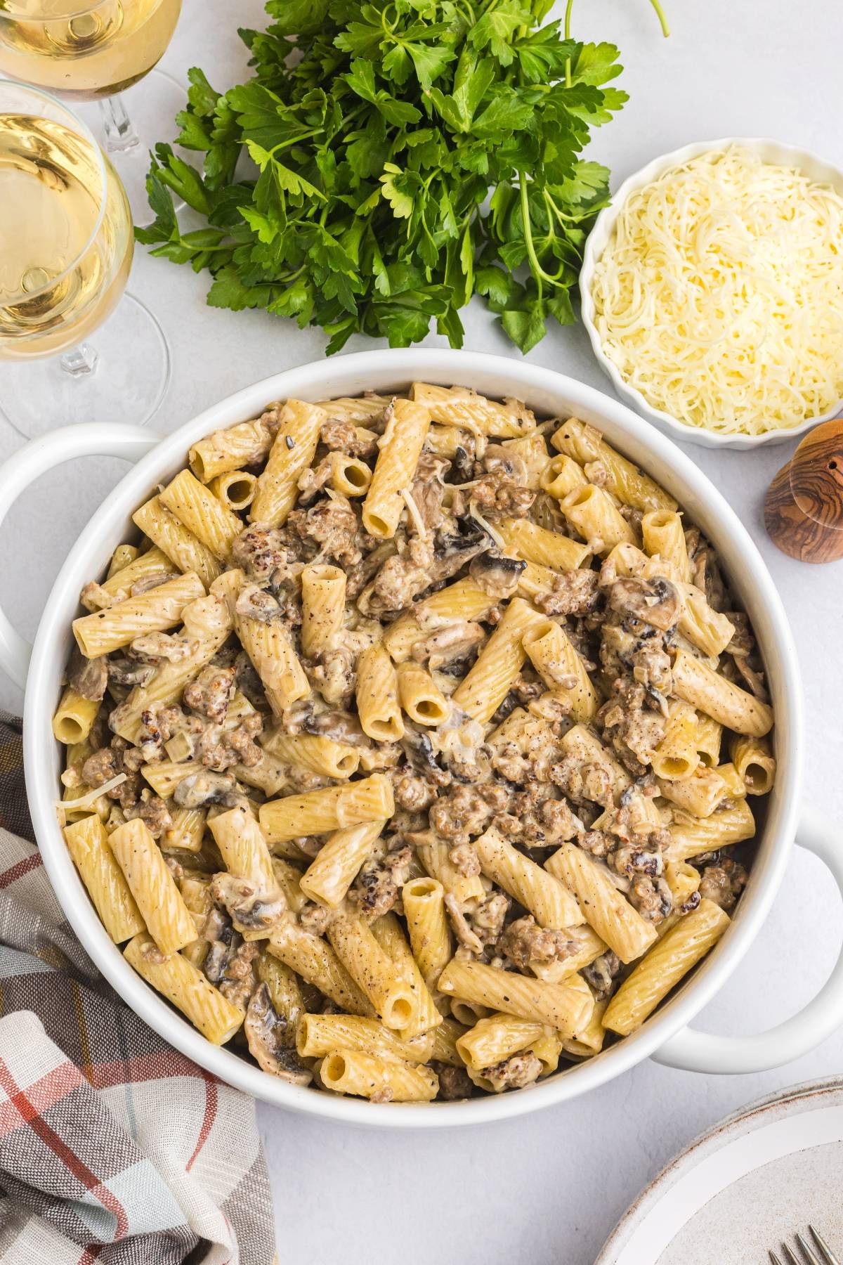 Creamy rigatoni pasta with ground beef and mushrooms in a white dish, surrounded by cheese and fresh parsley.