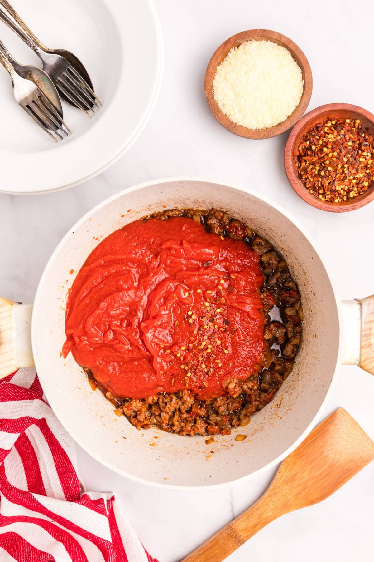 White pot with ground meat, tomato sauce, and chili flakes. Bowls of cheese and chili flakes and plates nearby.