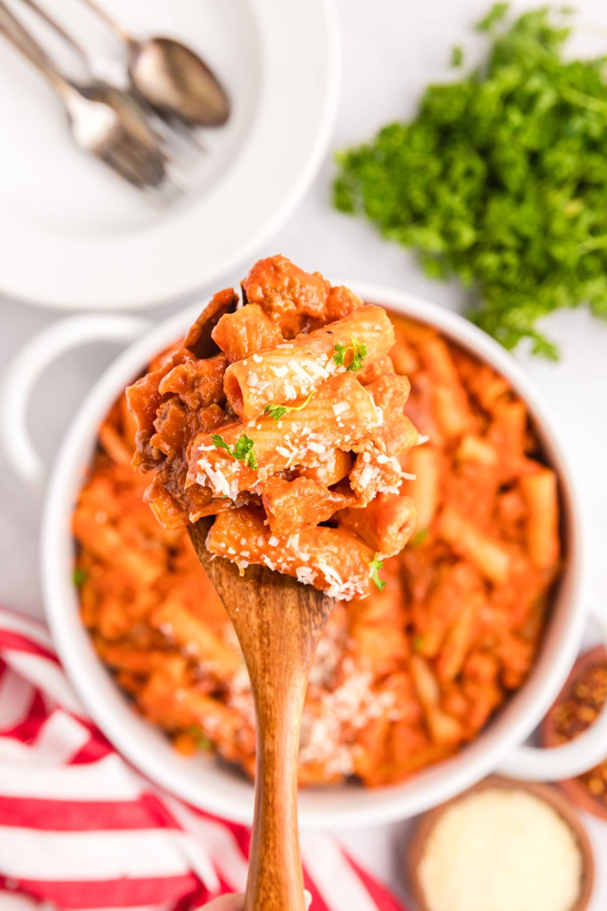 A wooden spoon lifts baked ziti with red sauce and herbs over a dish of pasta; parsley in background.