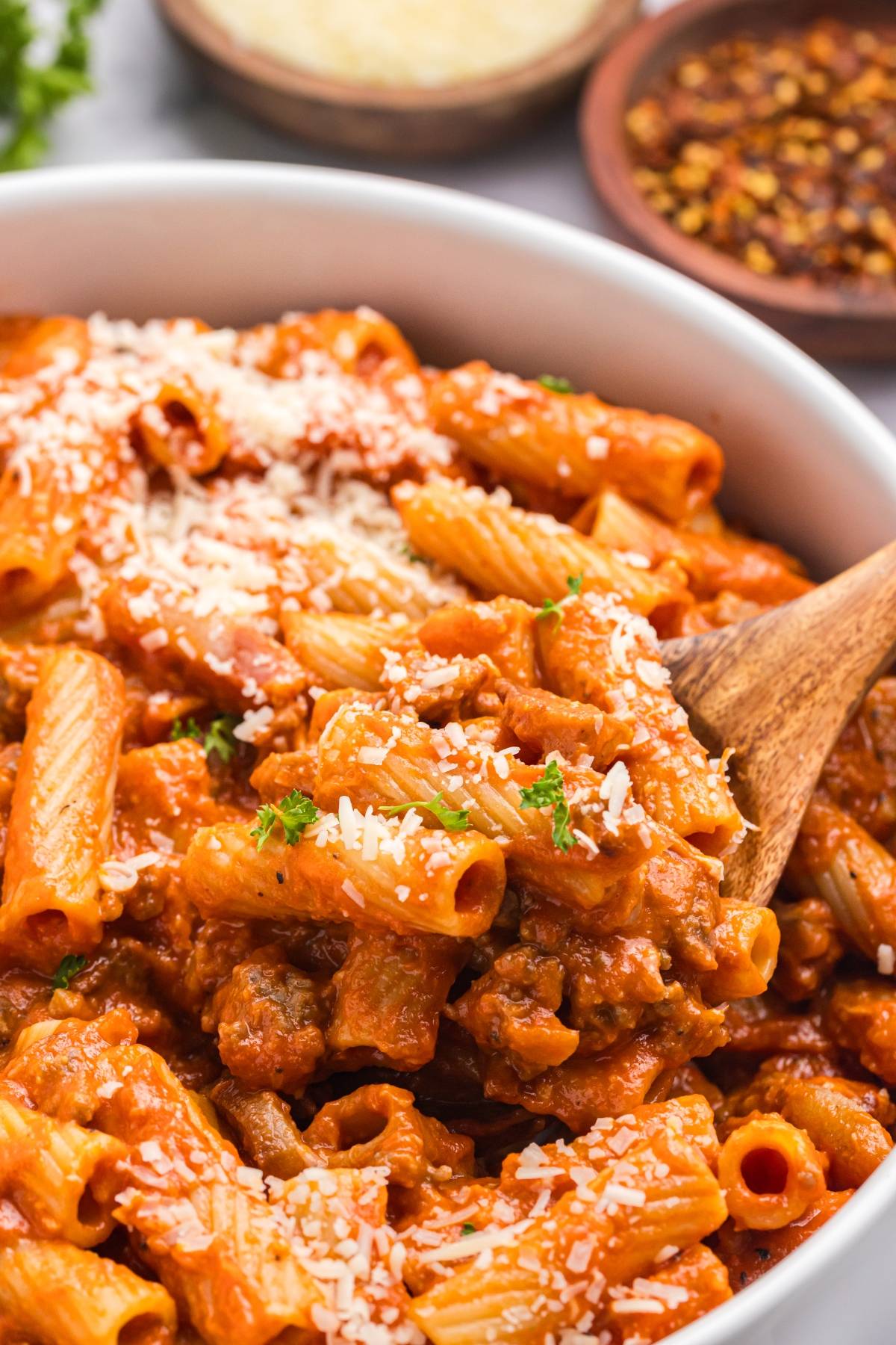 Close-up of baked rigatoni pasta with meat sauce, topped with grated cheese and parsley, served with a wooden spoon.