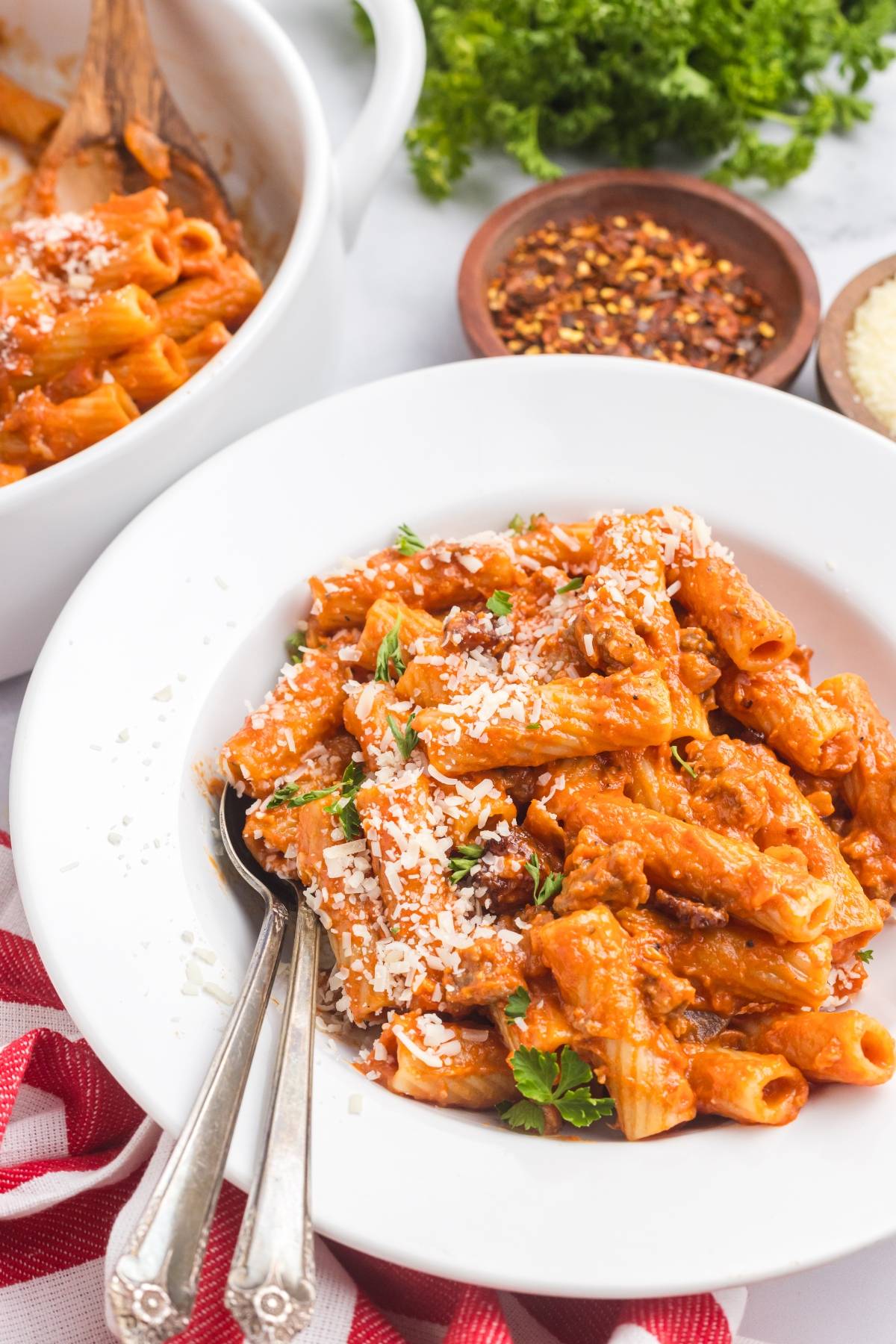 A white bowl of rigatoni pasta with meat sauce, topped with grated cheese and parsley, with two forks.