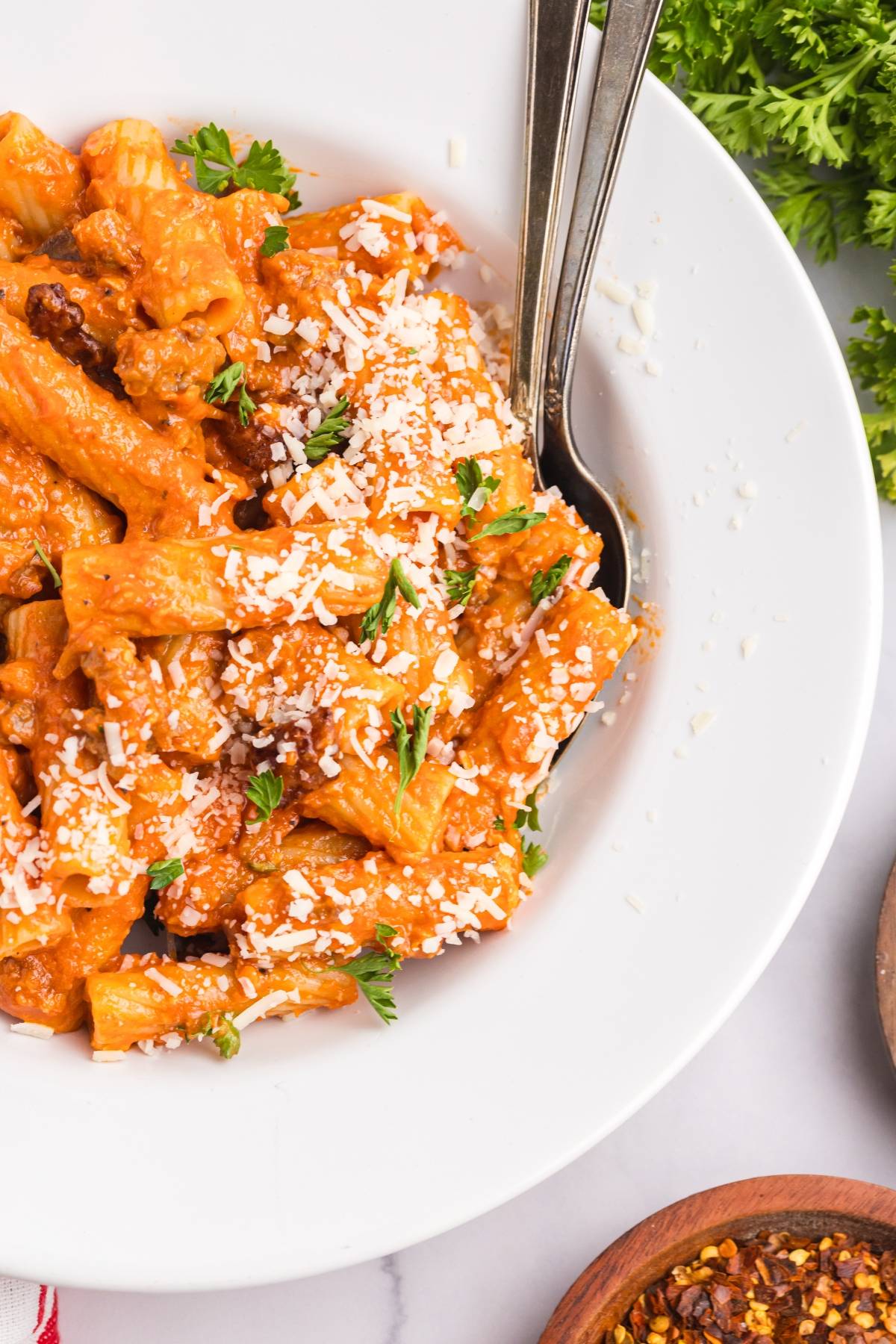 A white bowl of rigatoni pasta with tomato sauce, grated cheese, and parsley, with two forks beside.