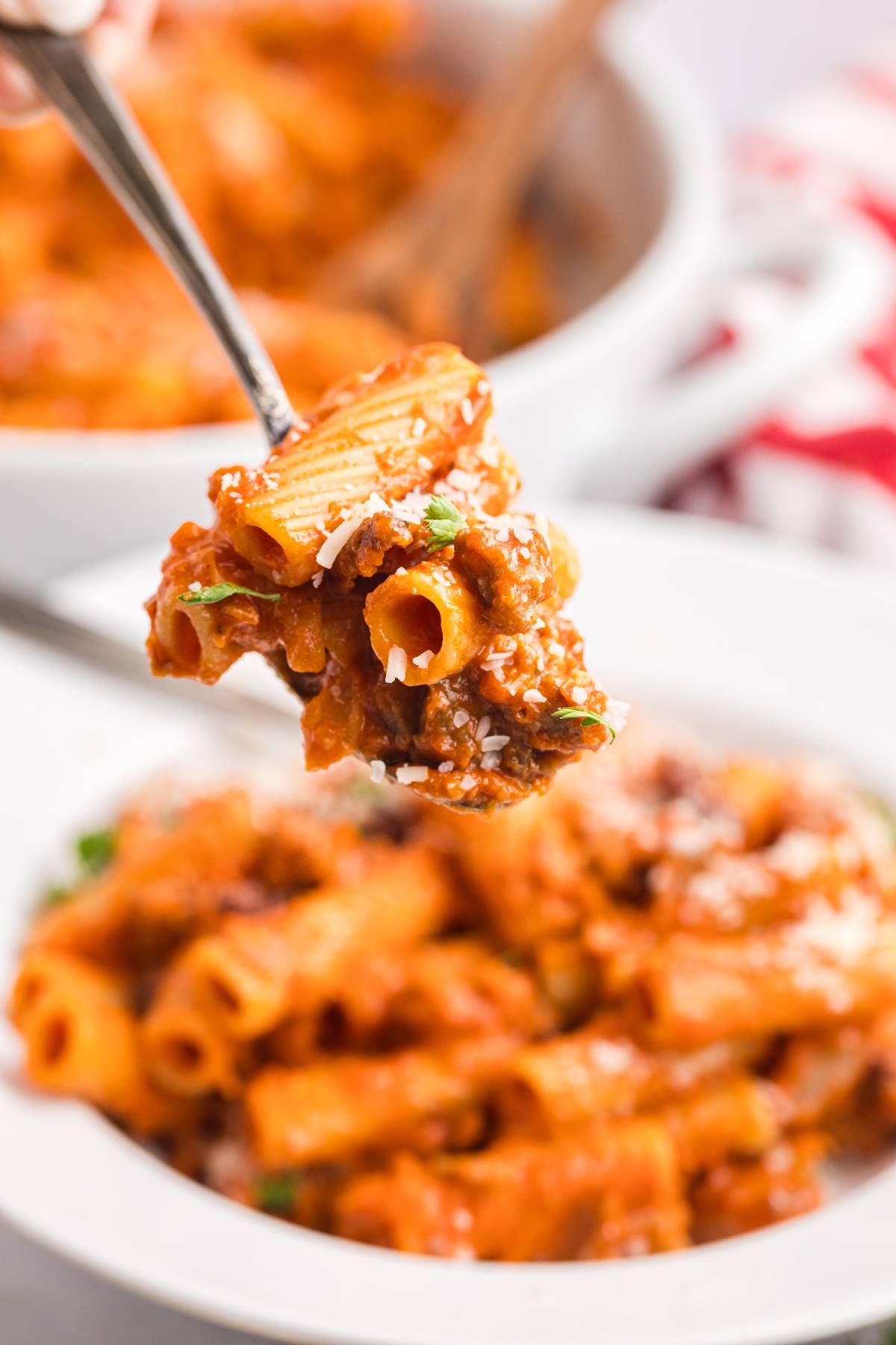 A fork holds up saucy rigatoni pasta with meat and herbs over a bowl of pasta.