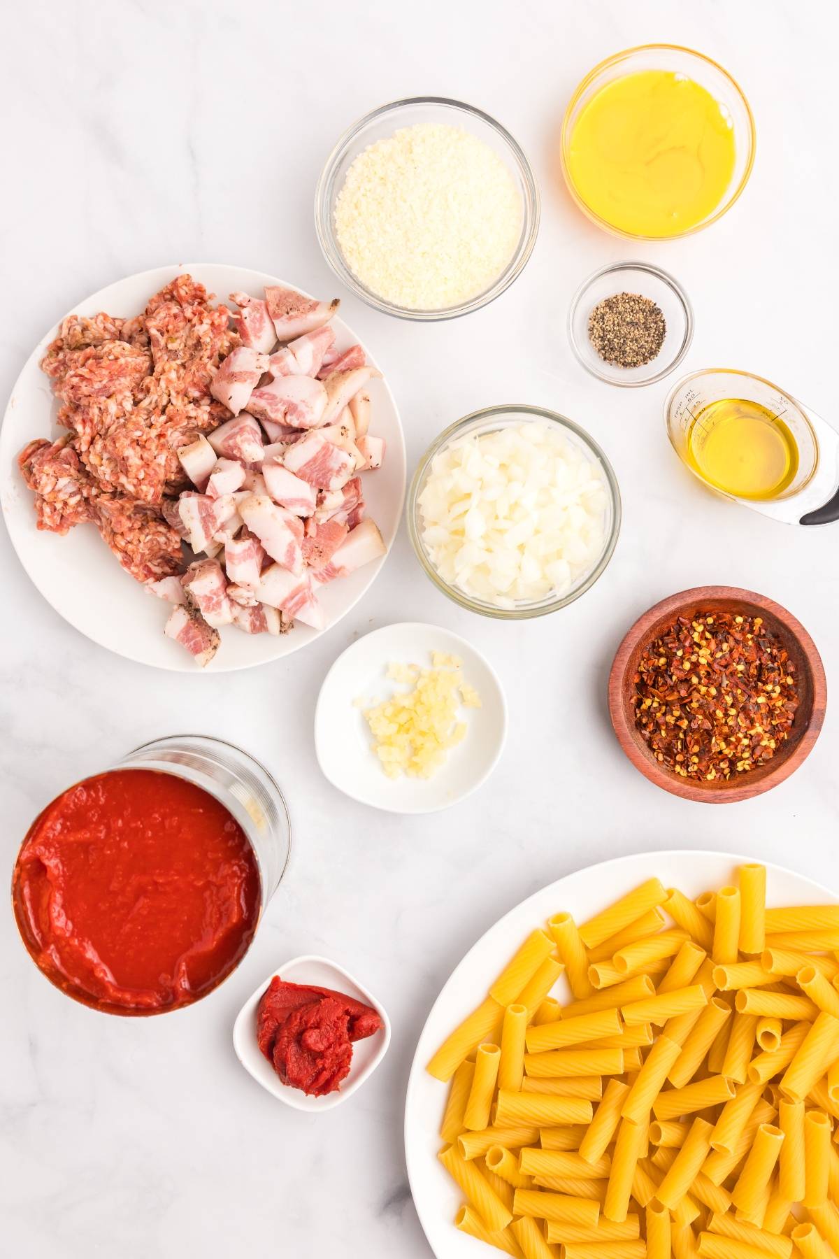 Various ingredients for pasta including raw sausage, bacon, rigatoni, tomato sauce, cheese, onions, and spices.