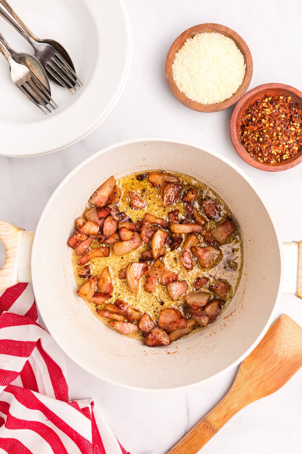A white pot with cooked bacon pieces in oil, next to bowls of cheese, chili flakes, and a red-striped towel.