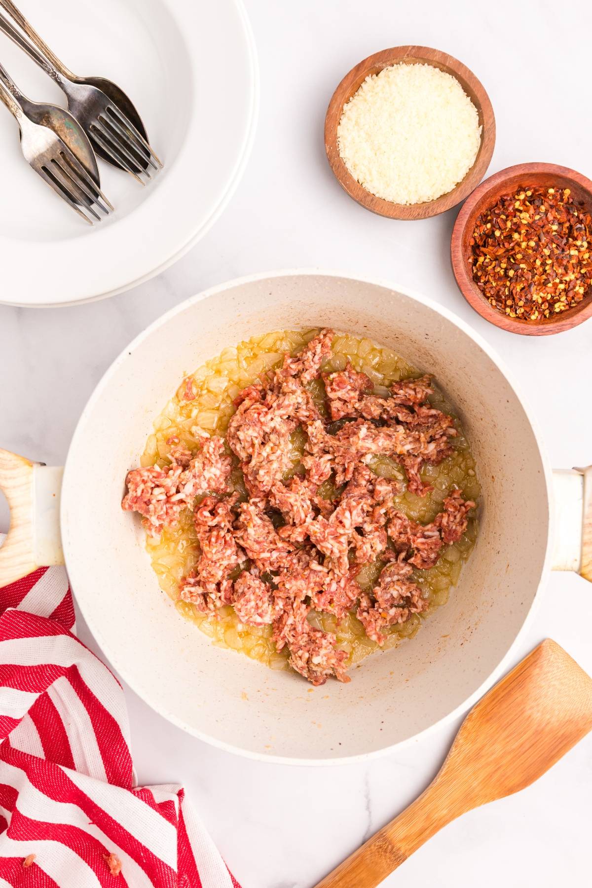 Ground meat cooking with onions in a white pan, surrounded by seasonings and utensils on a white surface.
