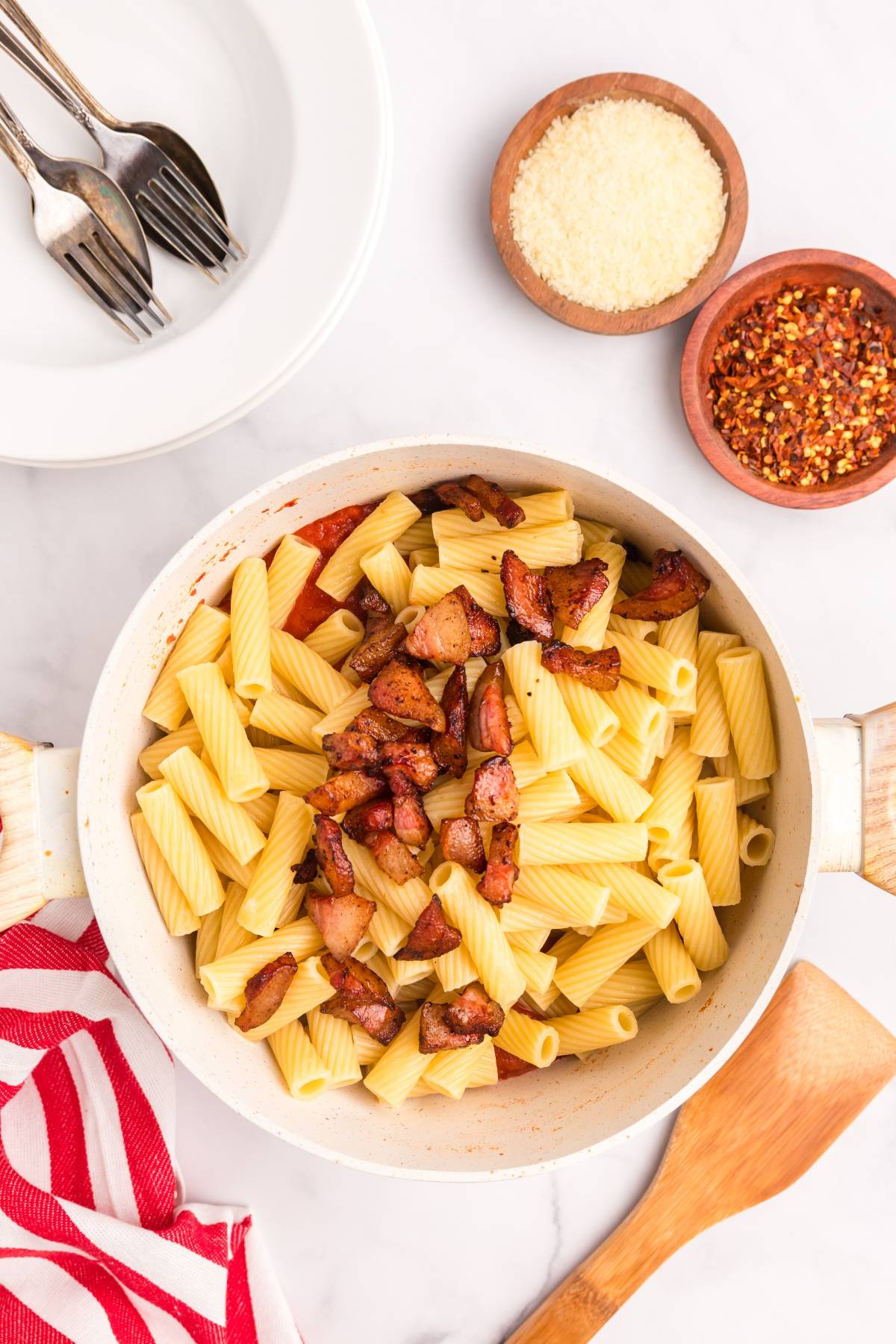 A pot of rigatoni pasta with crispy bacon, next to bowls of grated cheese and red pepper flakes.