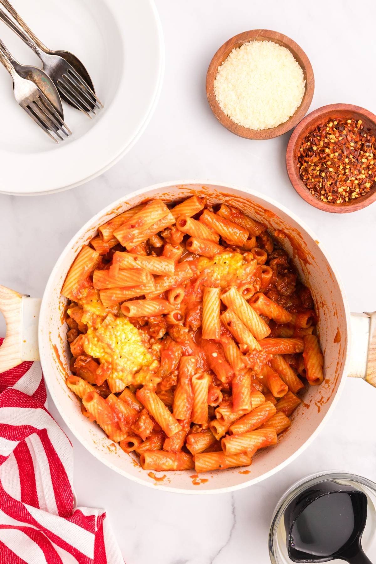 Pasta with tomato sauce and cheese in a pot, surrounded by parmesan, red pepper flakes, and tableware.