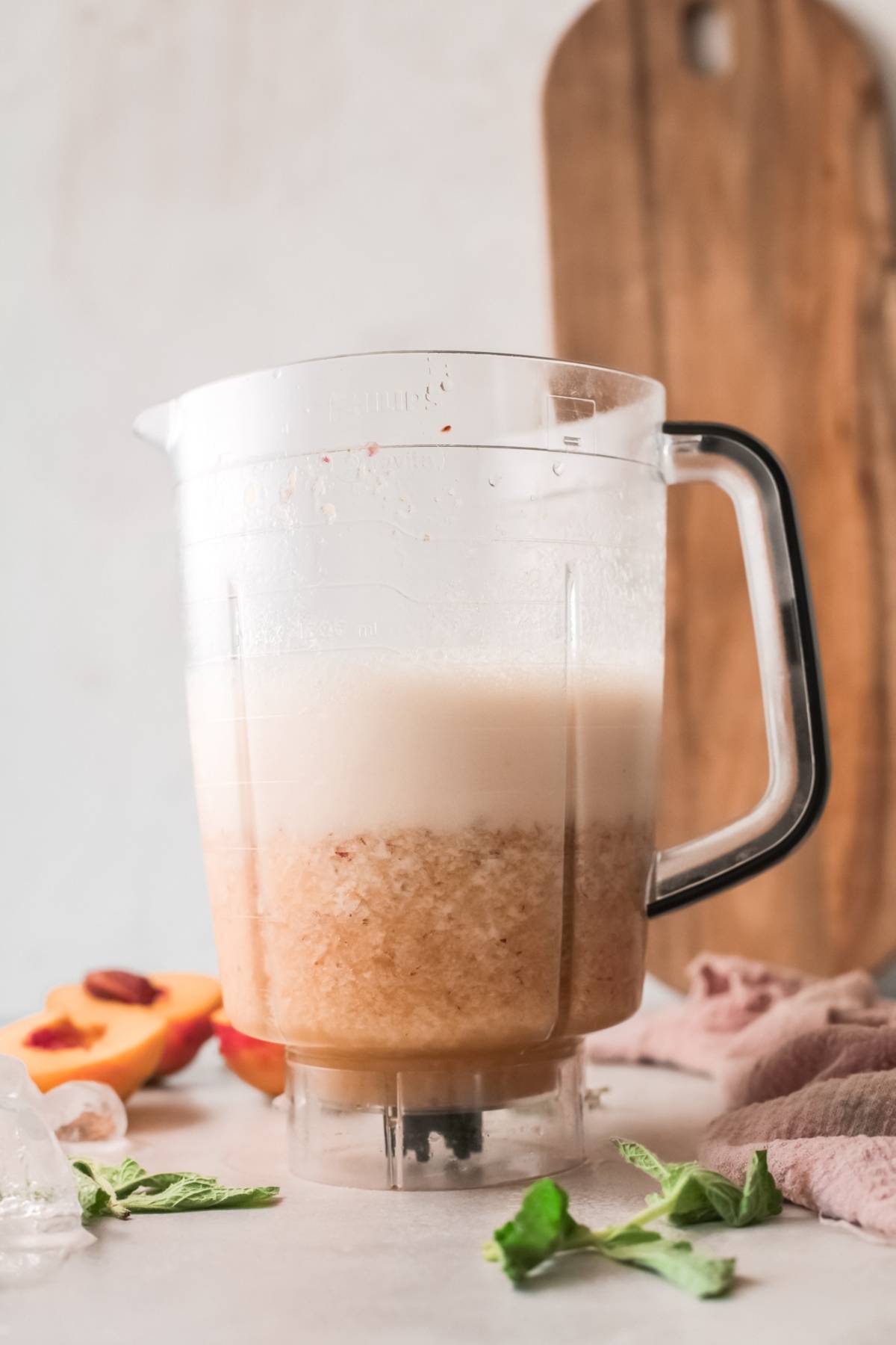 A blender pitcher with a frothy, light brown peach bellini smoothie sits next to ice, peach slices, mint, and a towel—perfect for those craving a Peach and prosecco twist.
