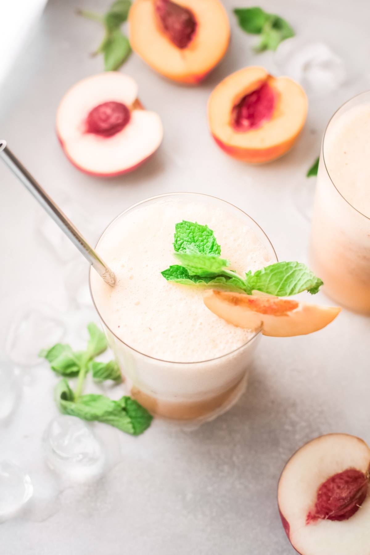 Iced peach smoothie garnished with mint and a peach slice, reminiscent of a refreshing peach bellini, served alongside fresh peach halves and ice.