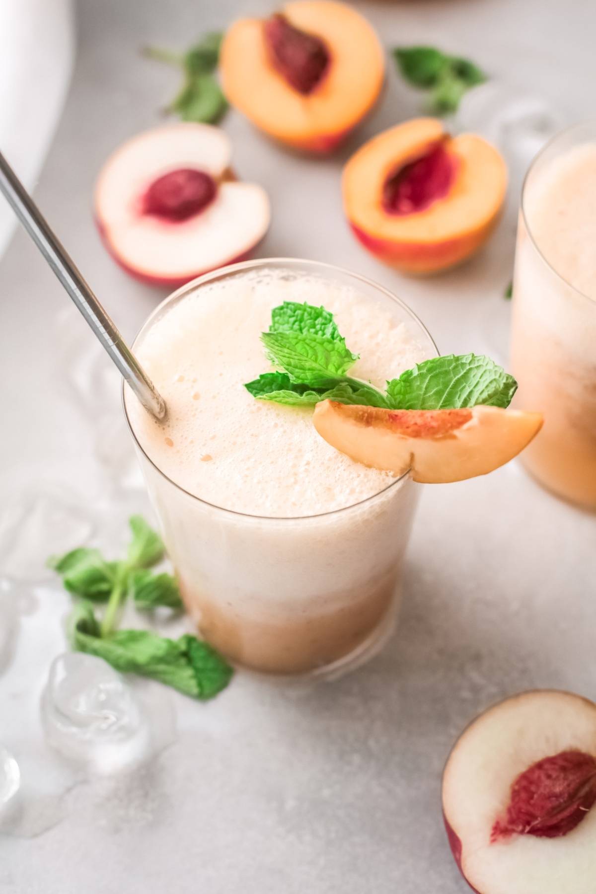 A peach smoothie in a glass with a metal straw, garnished with mint and a peach slice; fresh peaches nearby—like a refreshing recipe with peach juice, perfect for fans of Peach and prosecco.