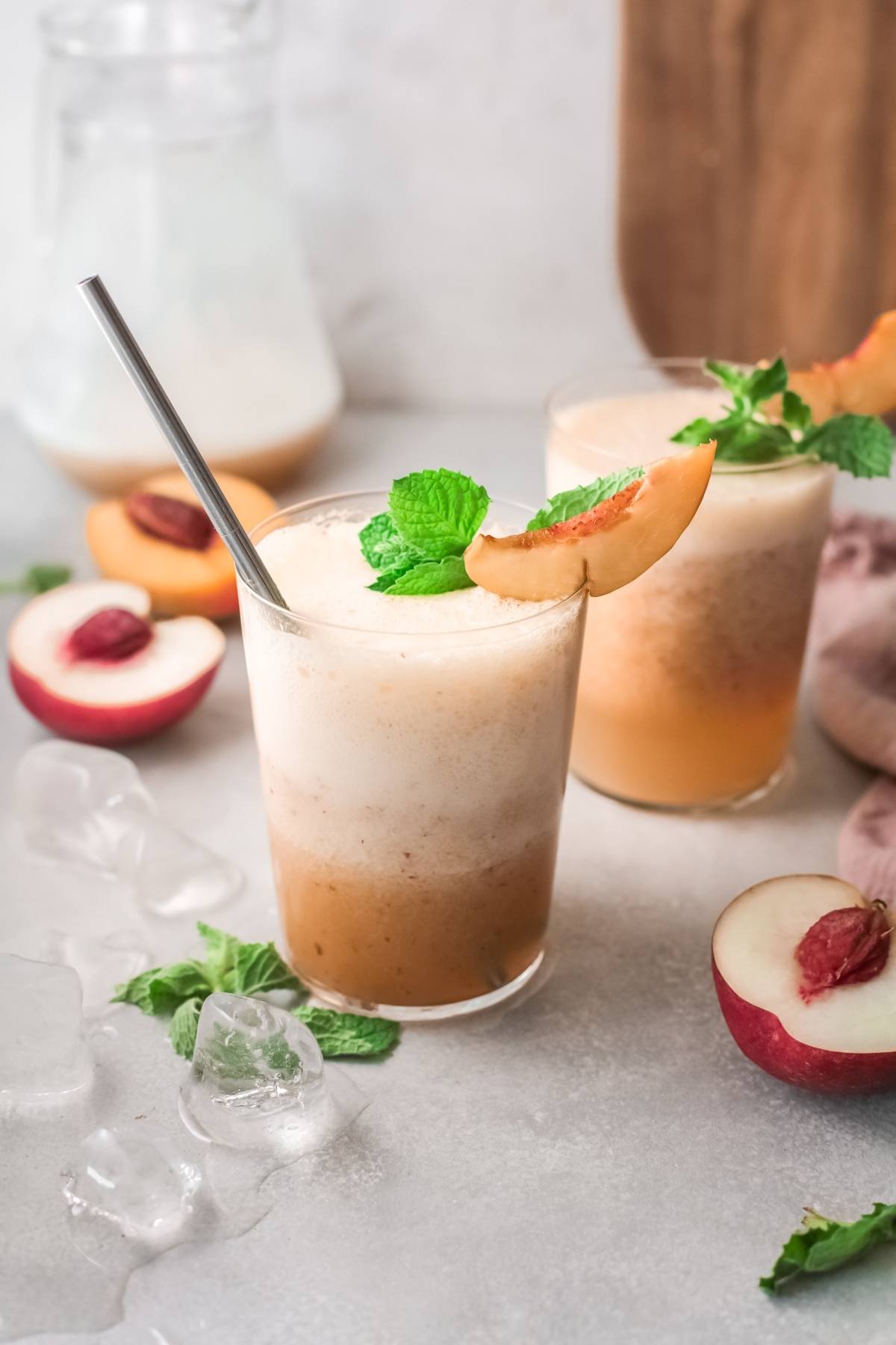 Two peach smoothies in glasses with metal straws, garnished with mint and peach slices, surrounded by ice and peaches—perfect inspiration for a refreshing recipe with peach juice or even a nonalcoholic twist on the classic Peach Bellini.