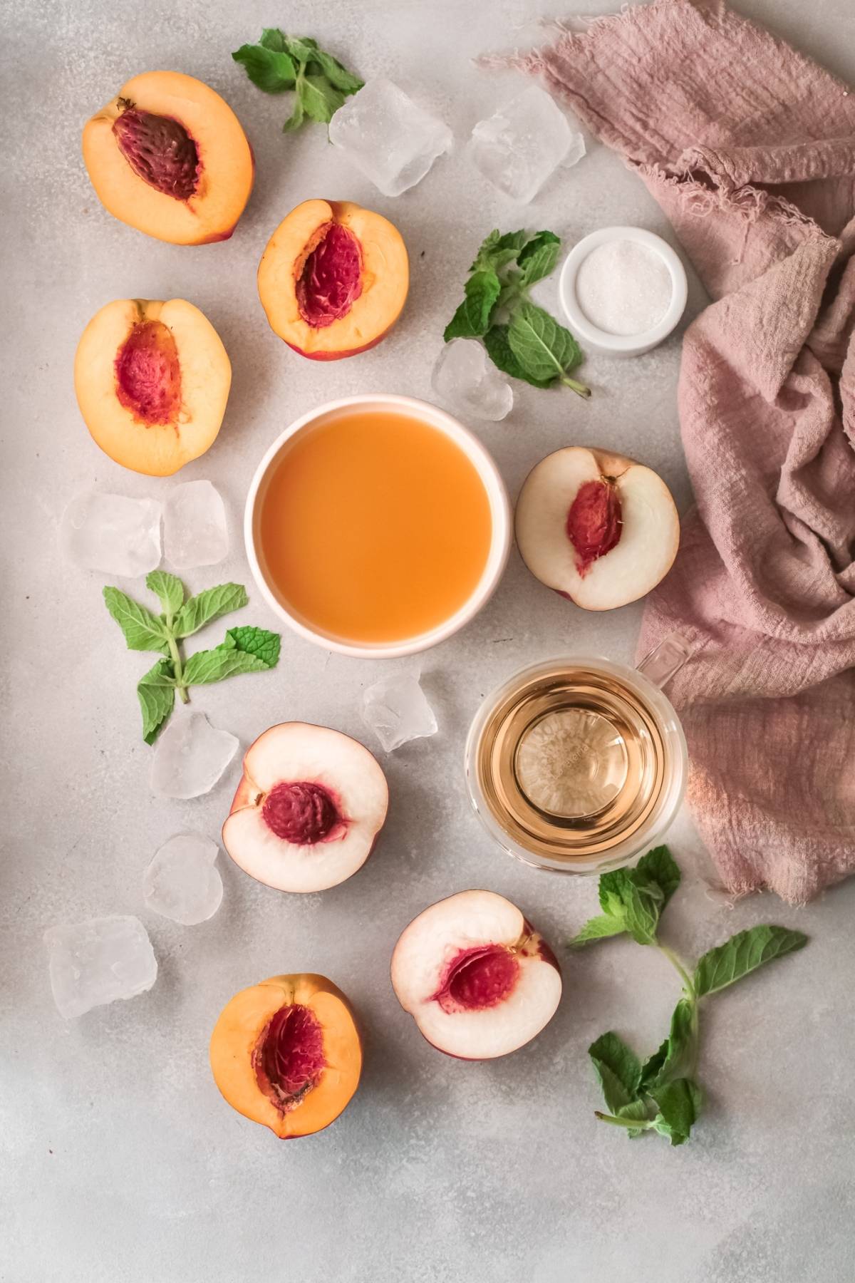 Peach halves, mint leaves, ice cubes, and peach juice evoke the refreshing ingredients of a classic Peach Bellini cocktail recipe, all artfully arranged on a beige cloth atop a light surface.