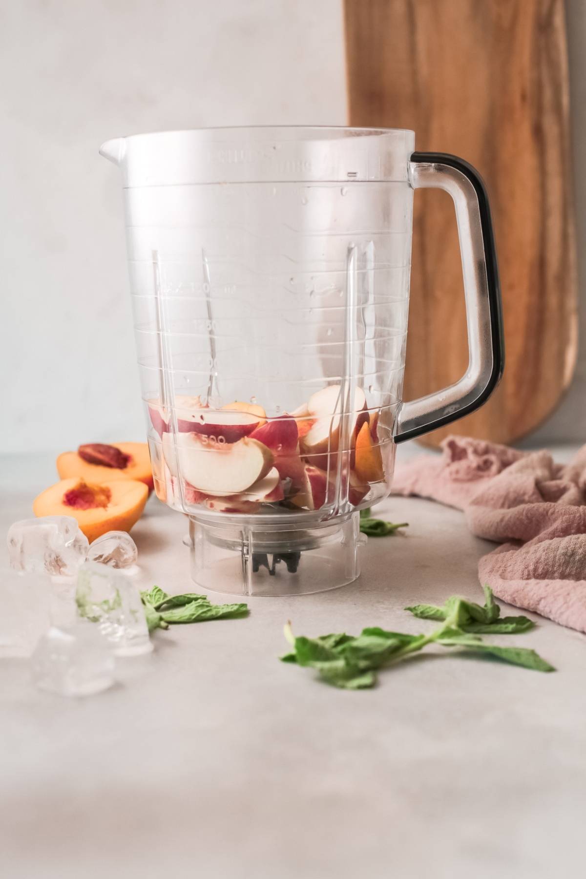 Blender pitcher with sliced peaches inside, perfect for a peach bellini, surrounded by ice cubes and fresh mint on a countertop.