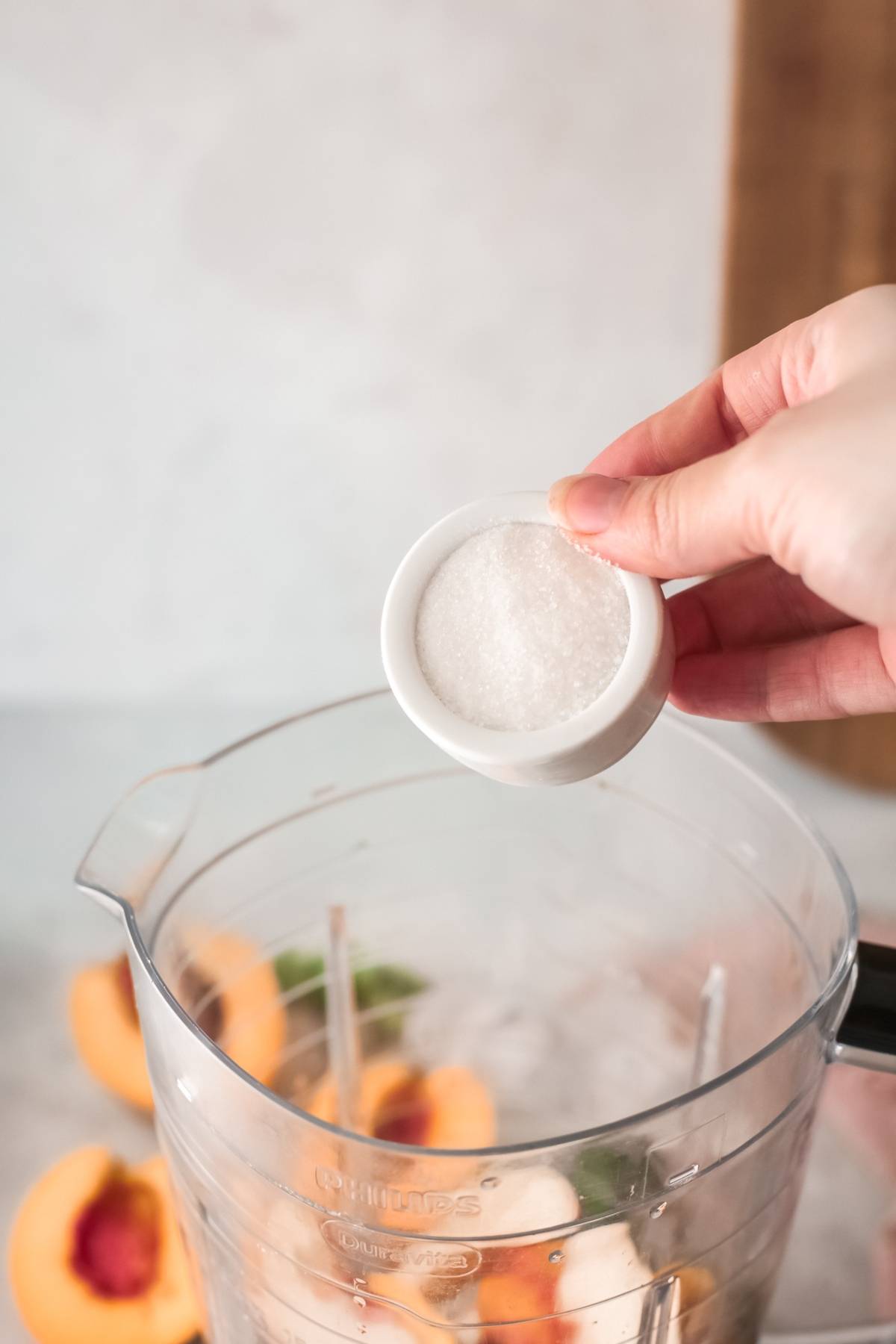 A hand holds a small bowl of sugar above a blender filled with peach halves and ice, just the start of a delicious Bellini recipe.