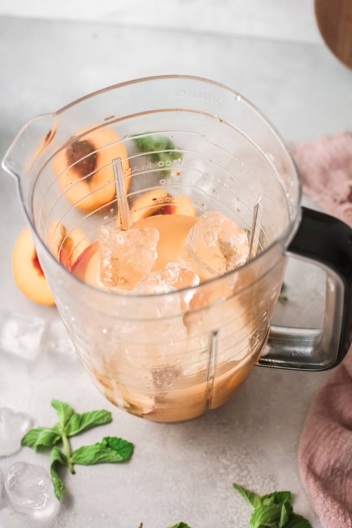 A blender pitcher filled with peach slices, ice cubes, and liquid, surrounded by mint leaves—perfect for trying a refreshing Bellini recipe.