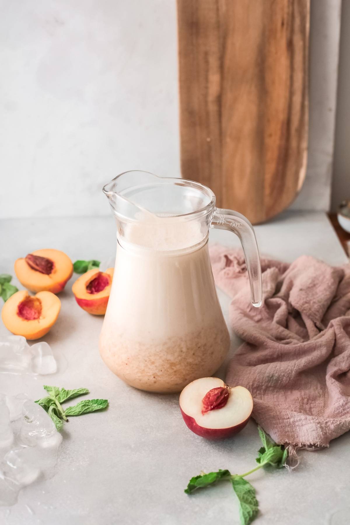 A glass pitcher of creamy peach bellini sits on a table with fresh peaches, ice cubes, and mint leaves nearby—perfect for those seeking a refreshing Peach and Prosecco treat.