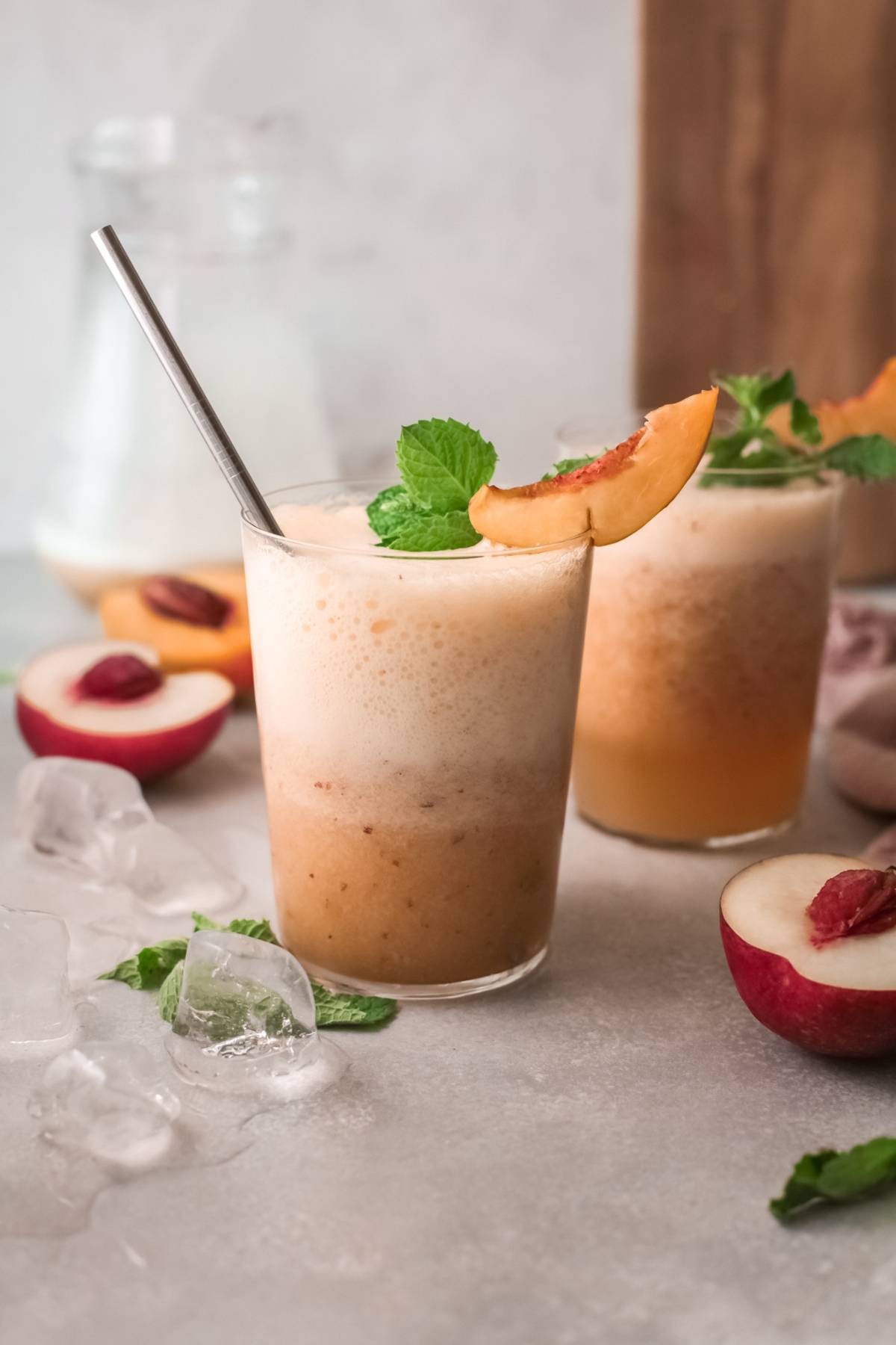 Iced peach smoothies in glasses, garnished with mint and peach slices, evoke the fresh sweetness of a peach bellini. Perfect for summer, this recipe with peach juice is served alongside ice and juicy peach halves.