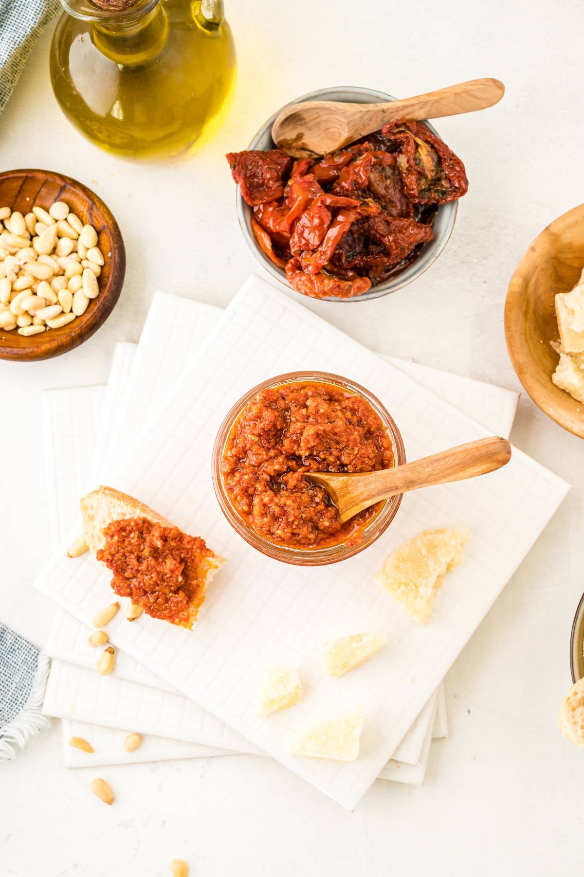 A jar of sun dried tomato pesto with a spoon, surrounded by bread, pine nuts, sun-dried tomatoes, and olive oil—a perfect red pesto recipe idea.