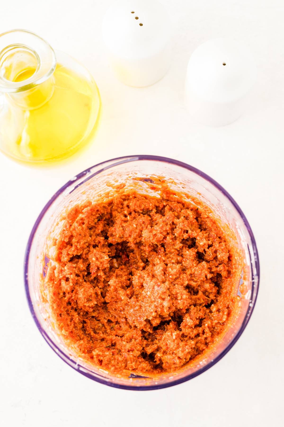 Food processor bowl with homemade Sun dried tomato pesto, next to a jar of olive oil and two white salt and pepper shakers.