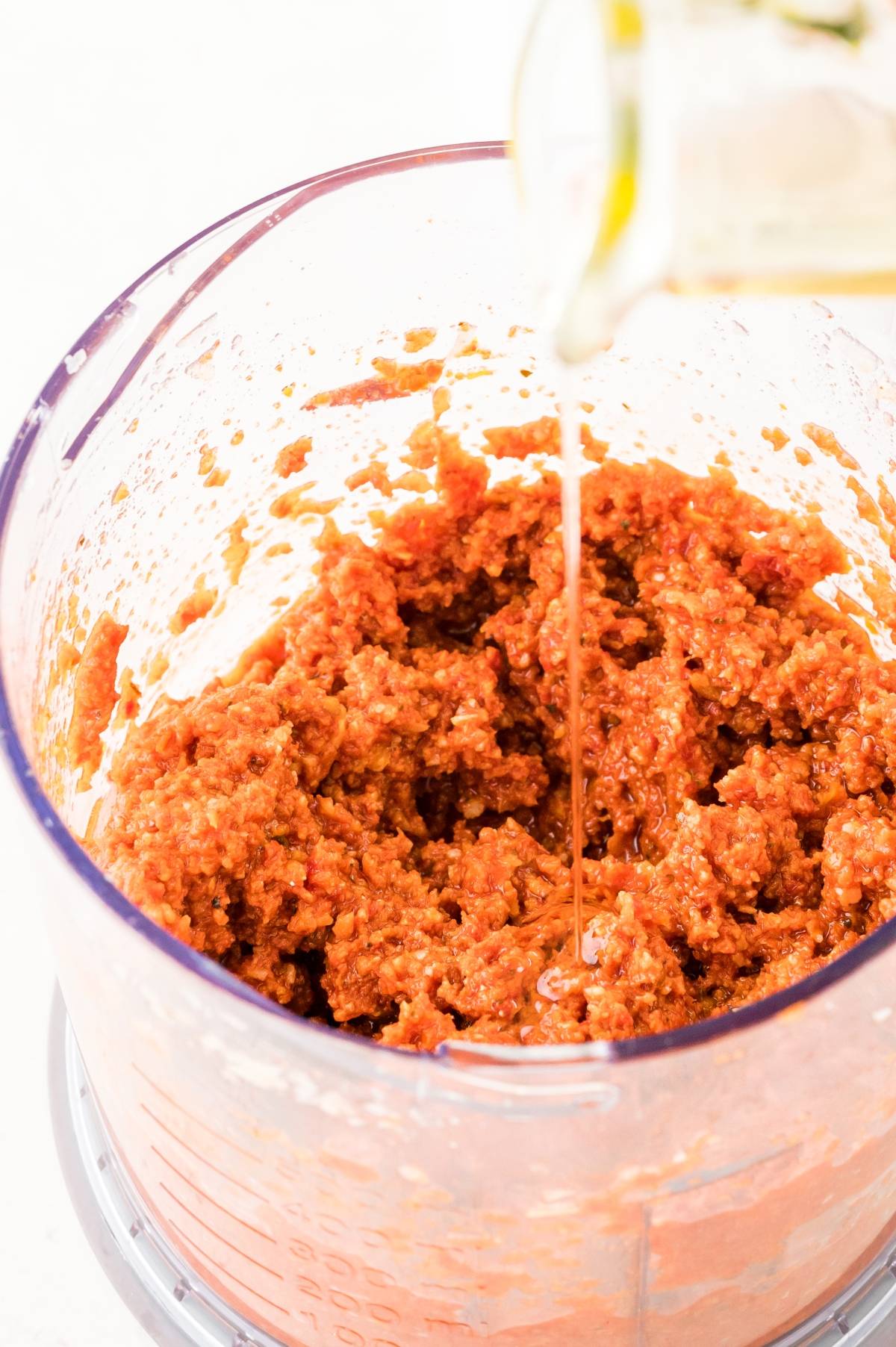 A vibrant red pesto recipe blends in a food processor as oil is poured from the top right, creating an easy pasta sauce packed with flavor.