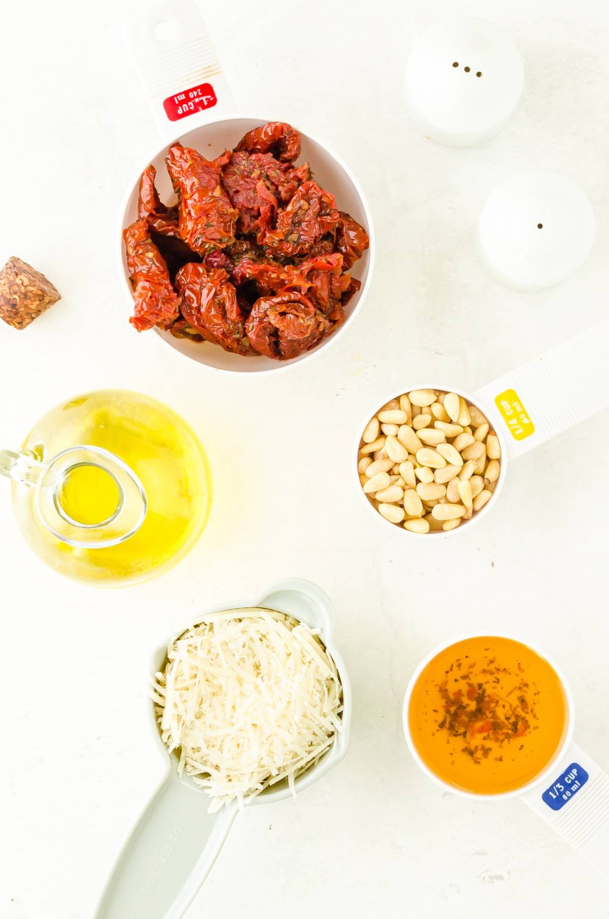 Measured ingredients for sun dried tomato pesto—sun-dried tomatoes, pine nuts, Parmesan, olive oil, and seasoning—are displayed on a white background.