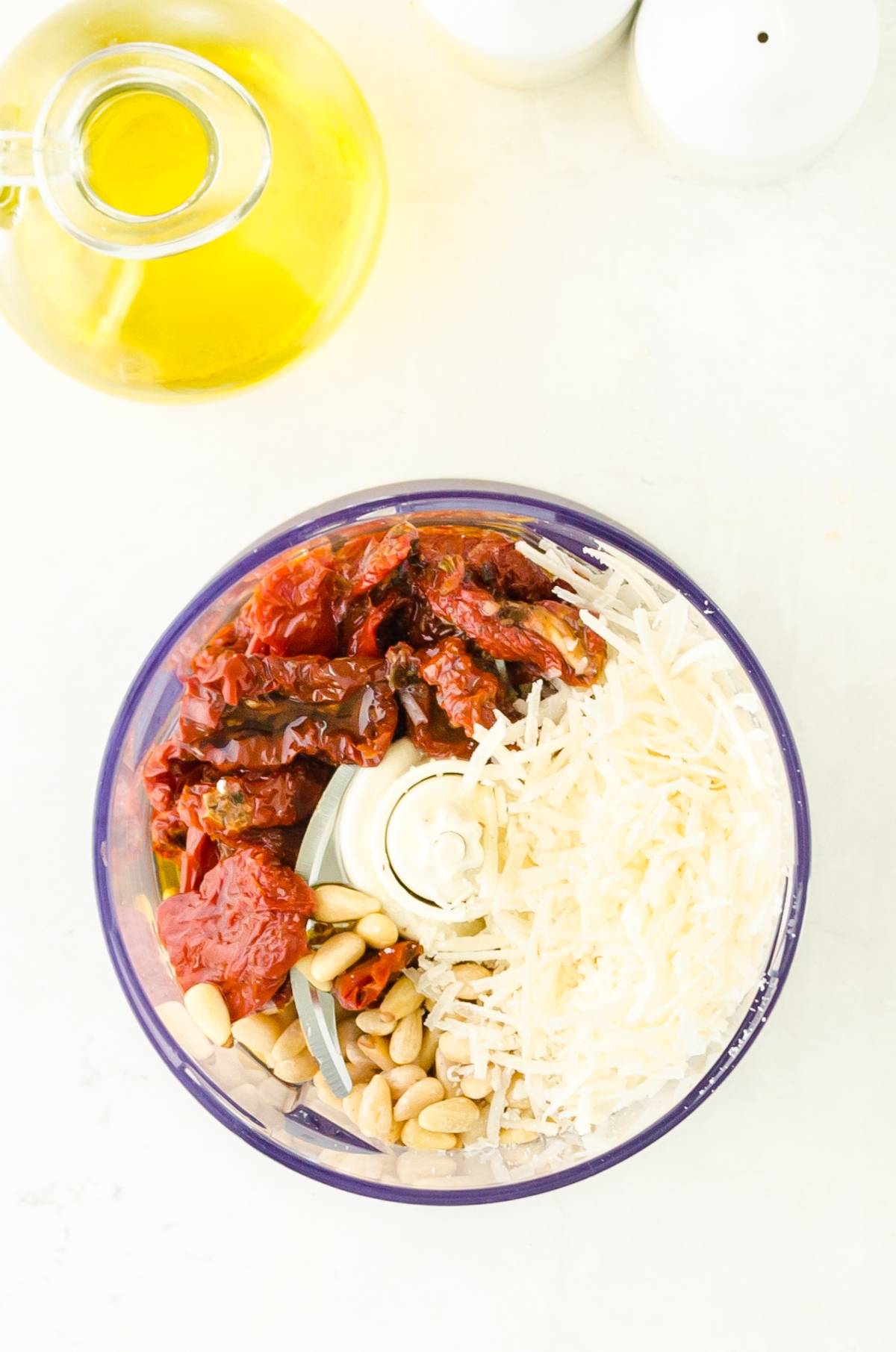 Food processor bowl filled with sun-dried tomatoes, grated cheese, pine nuts, and garlic; olive oil nearby—perfect for an Easy pasta sauce like Pesto Rosso – Sun Dried Tomato Pesto.