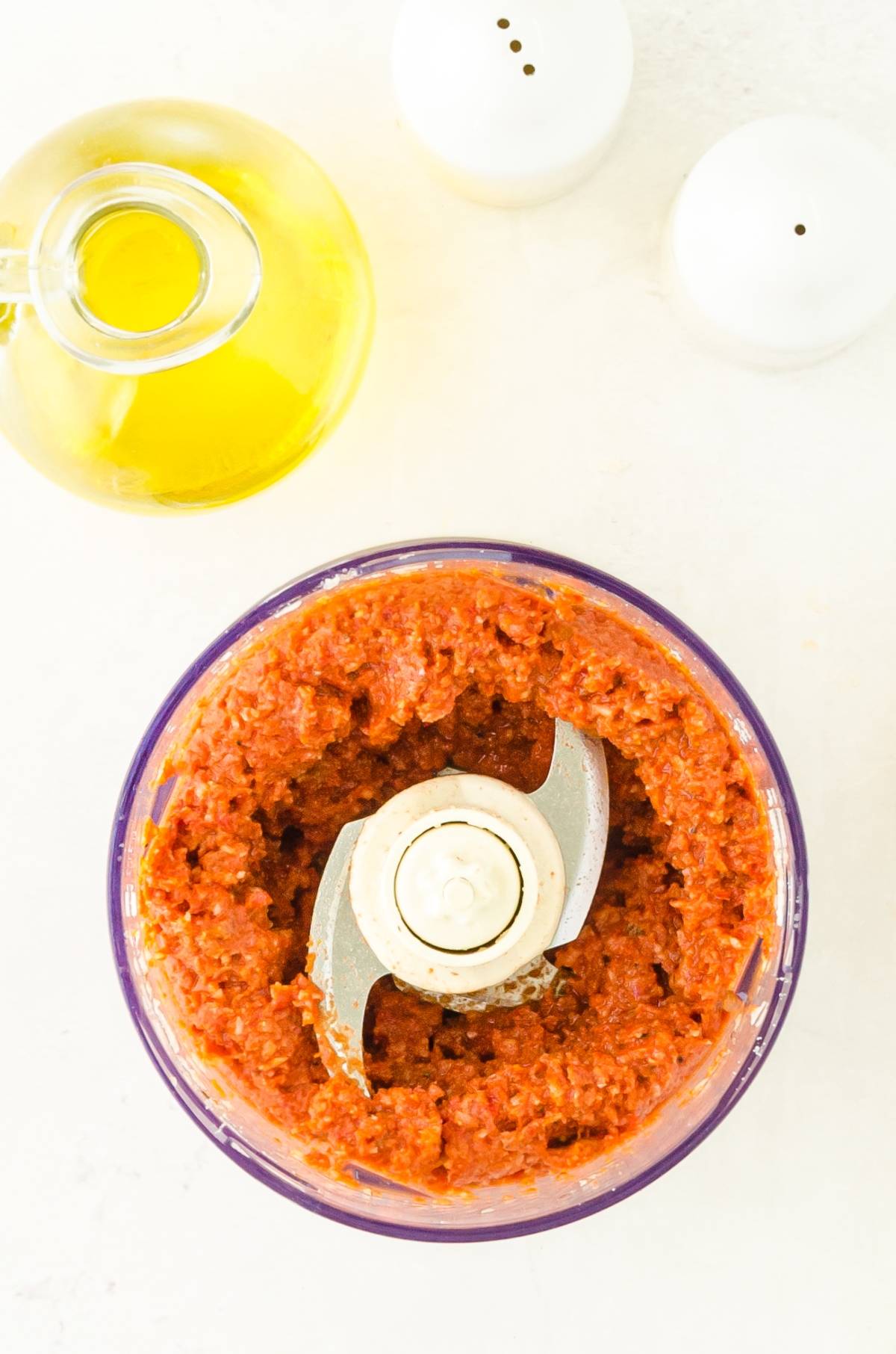 Top view of a food processor with blended Pesto Rosso – Sun Dried Tomato Pesto, olive oil, and two white shakers on a white surface.