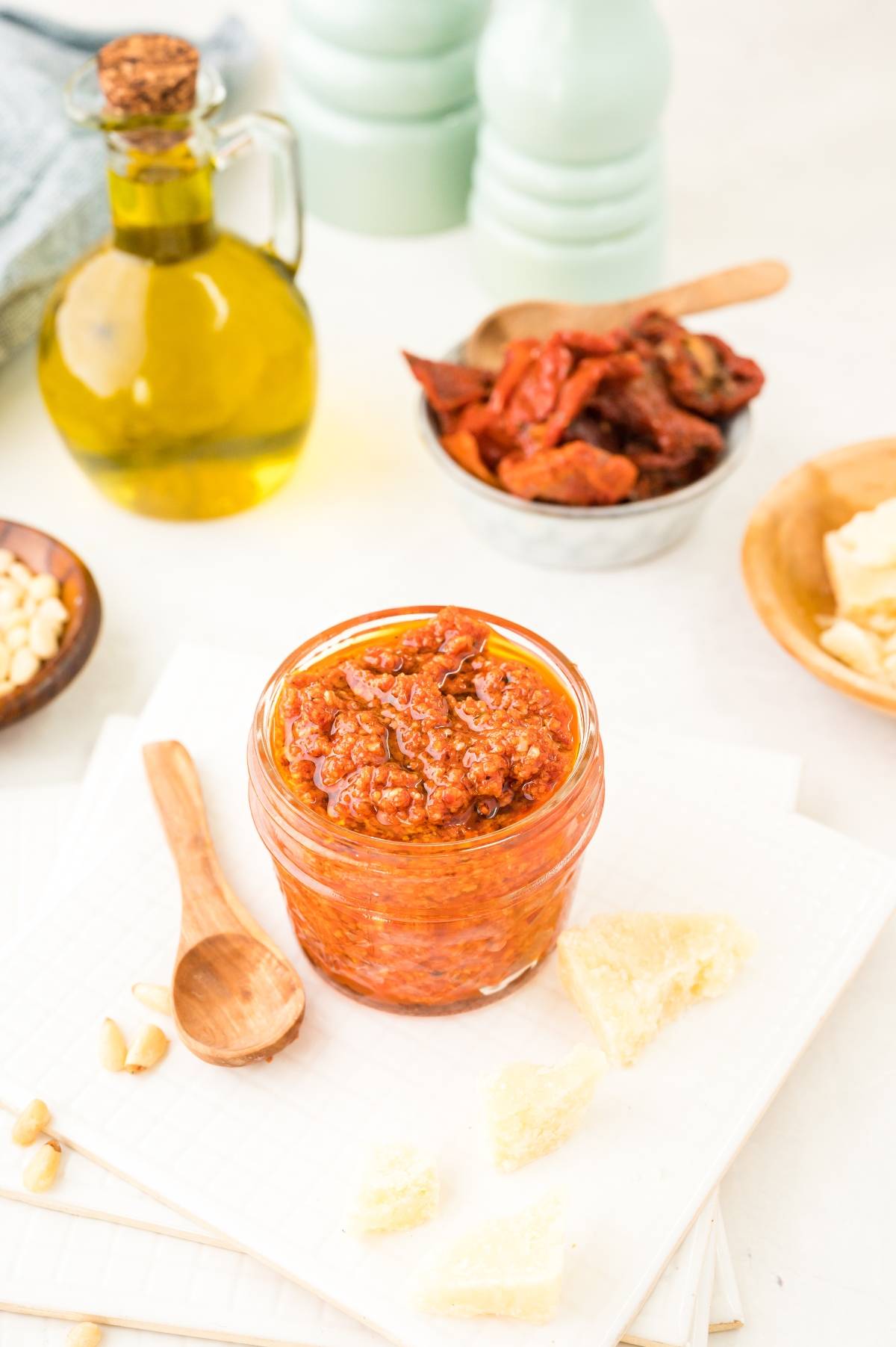 A jar of sun dried tomato pesto sauce surrounded by parmesan, pine nuts, dried tomatoes, and olive oil on a white surface—an easy pasta sauce perfect for any red pesto recipe.