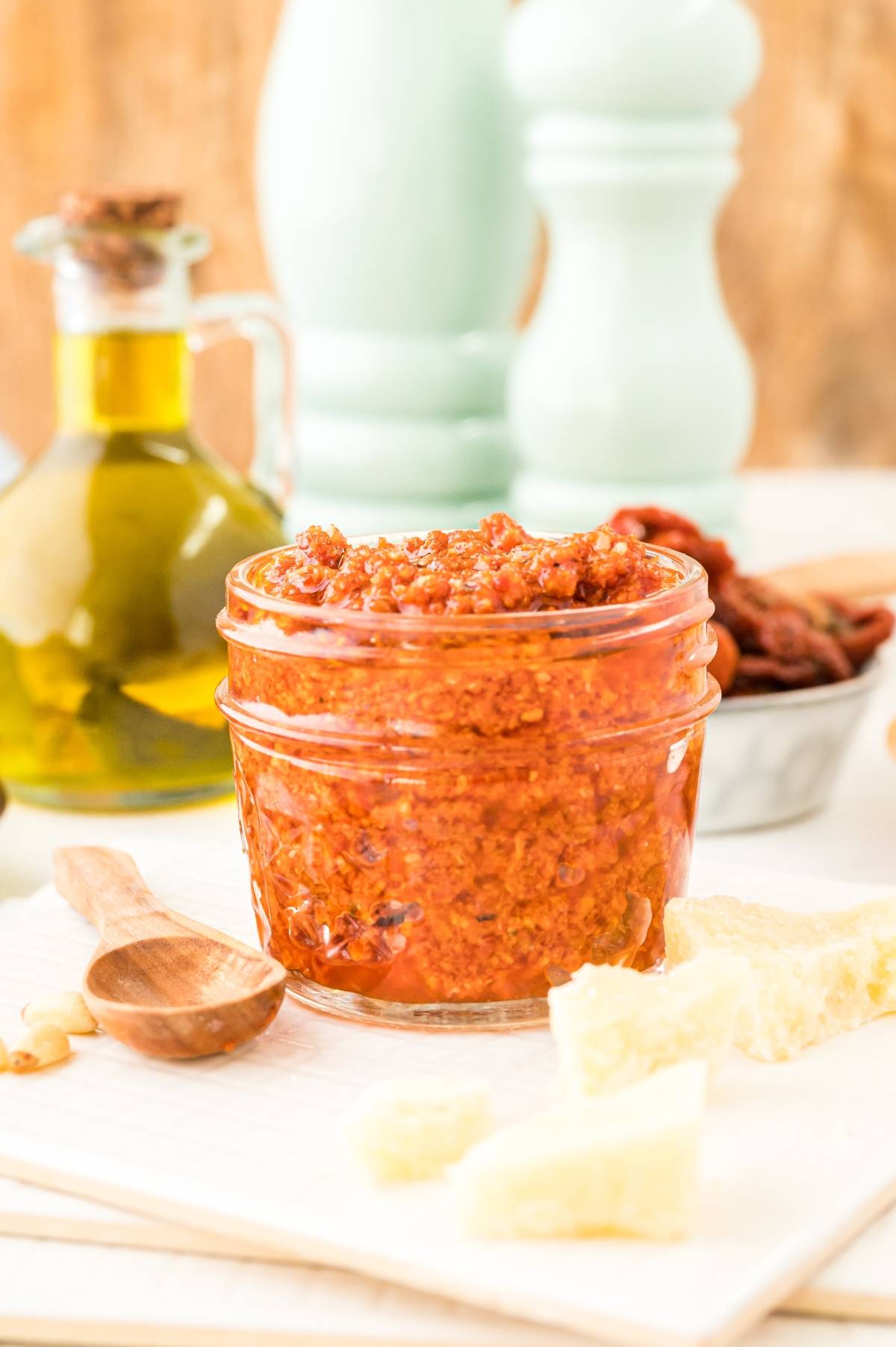A jar of sun dried tomato pesto with a wooden spoon, cheese chunks, and a bottle of olive oil on a table—an easy pasta sauce perfect for quick meals.
