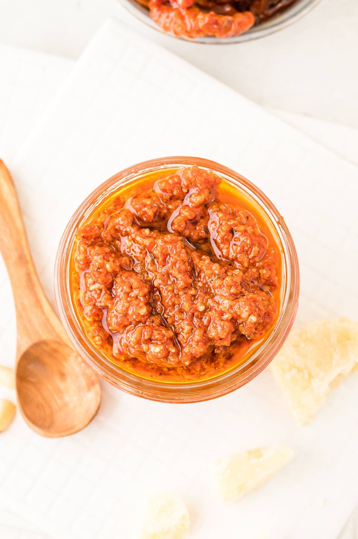 A jar of sun-dried tomato pesto sits on a white surface next to a wooden spoon and cheese pieces, ready to be enjoyed as an easy pasta sauce or used in your favorite red pesto recipe.
