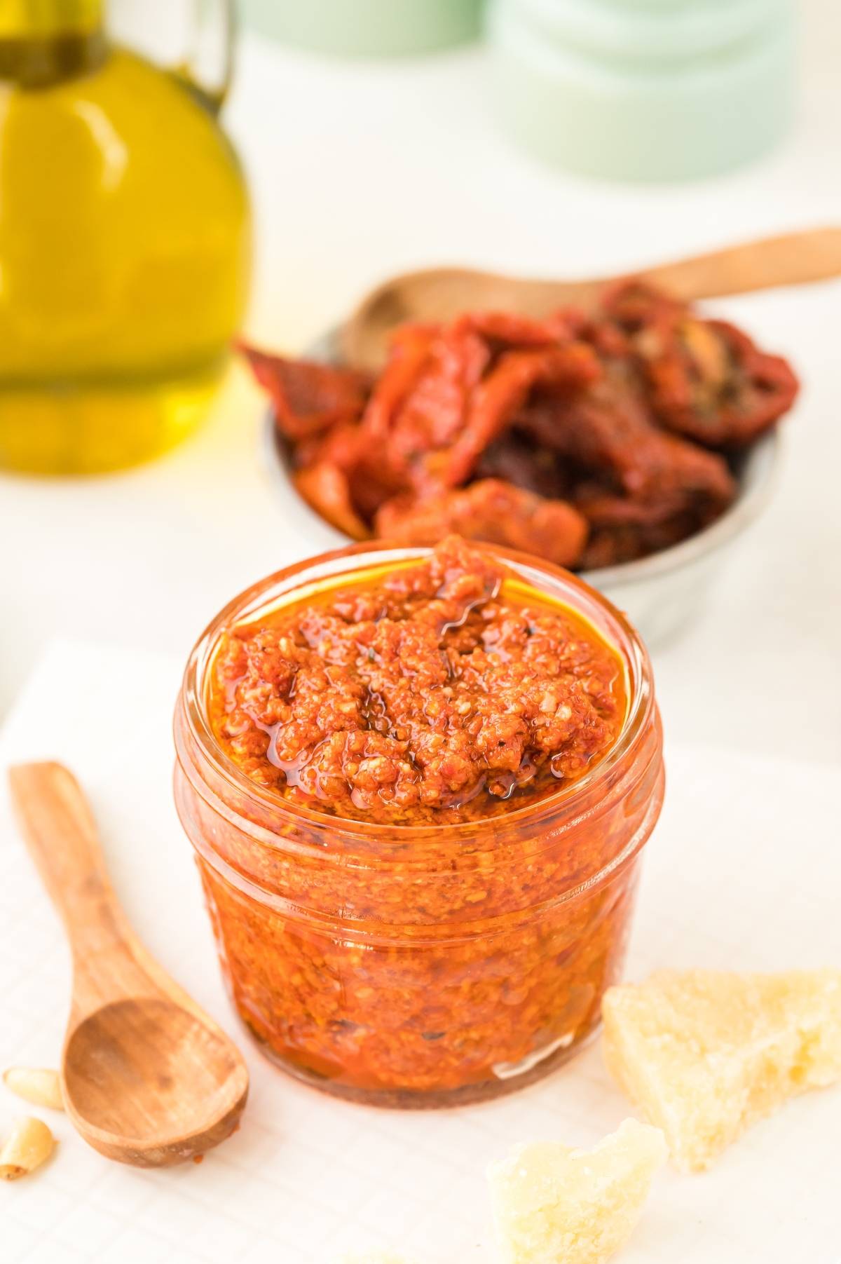 Small jar of Pesto Rosso – Sun Dried Tomato Pesto with a wooden spoon, cheese chunks, and olive oil in the background—perfect as an easy pasta sauce or for any recipe with sun dried tomatoes.
