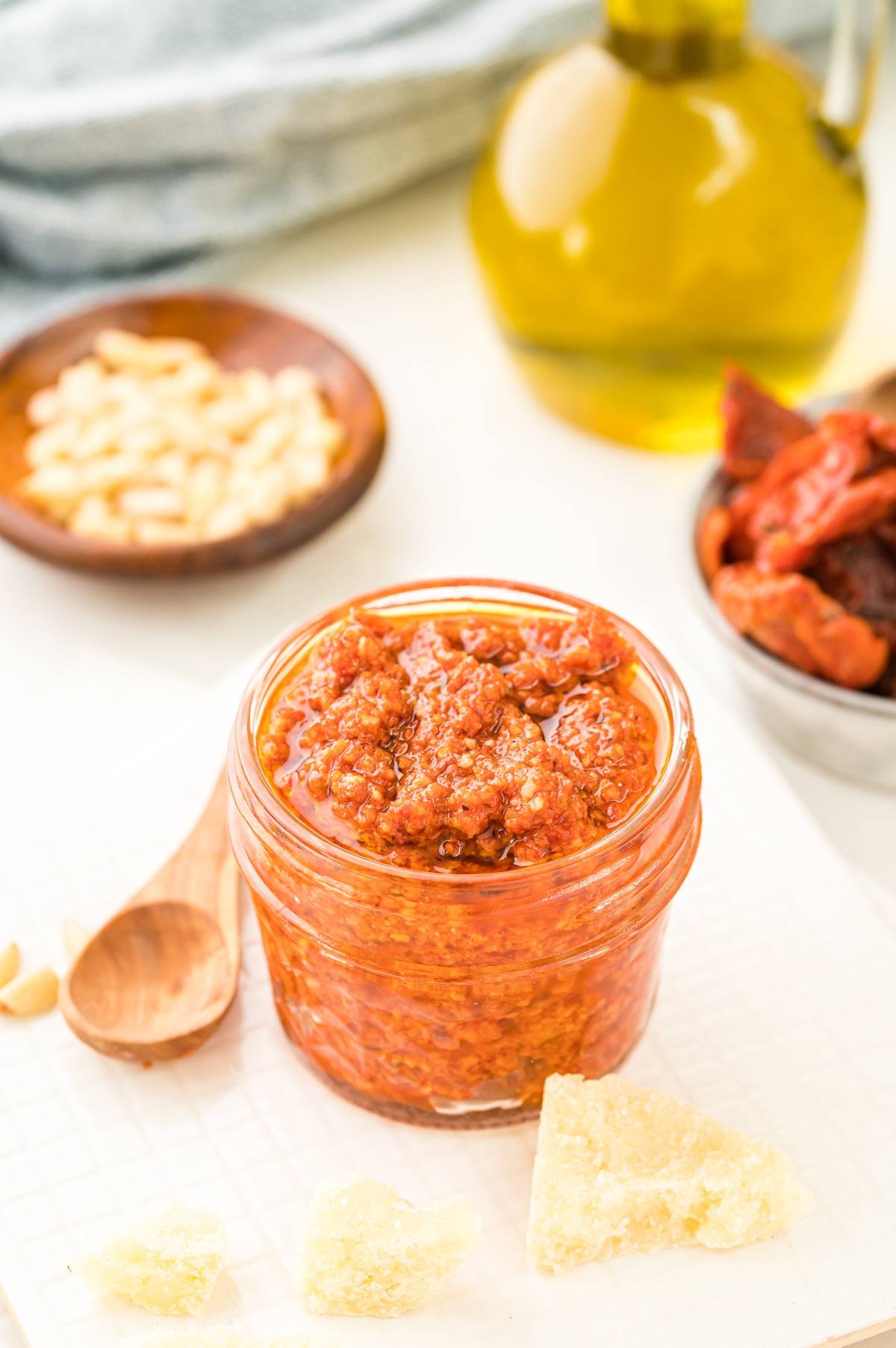 A small jar of Pesto Rosso – Sun Dried Tomato Pesto with a wooden spoon, cheese chunks, and fresh ingredients in the background.