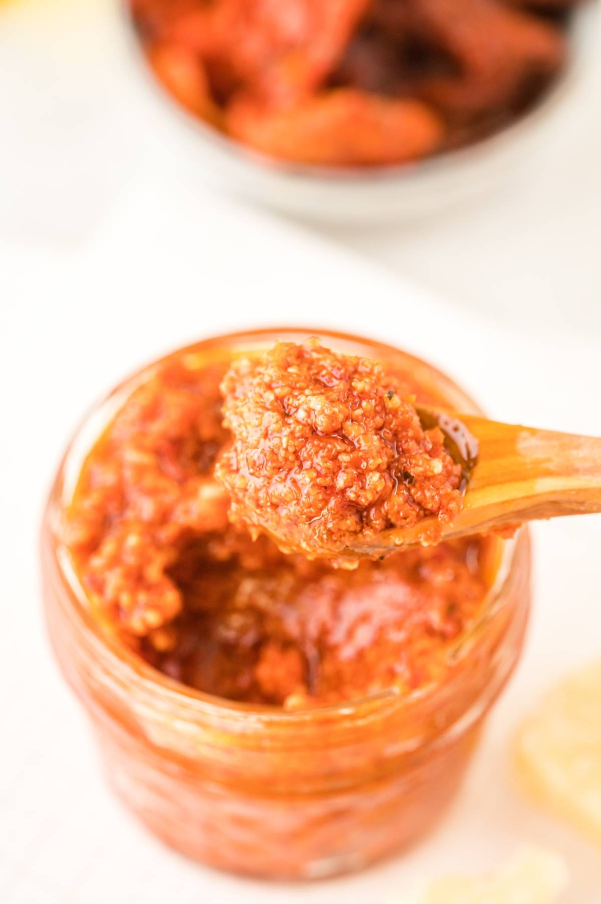 A wooden spoon scooping vibrant Pesto Rosso – Sun Dried Tomato Pesto from a glass jar, with a blurred bowl in the background—an easy pasta sauce perfect for your next red pesto recipe.