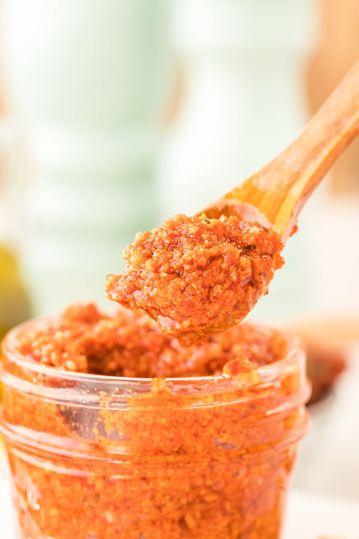 A wooden spoon scoops red pesto sauce from a small glass jar—an easy pasta sauce perfect for any recipe with sun dried tomatoes.