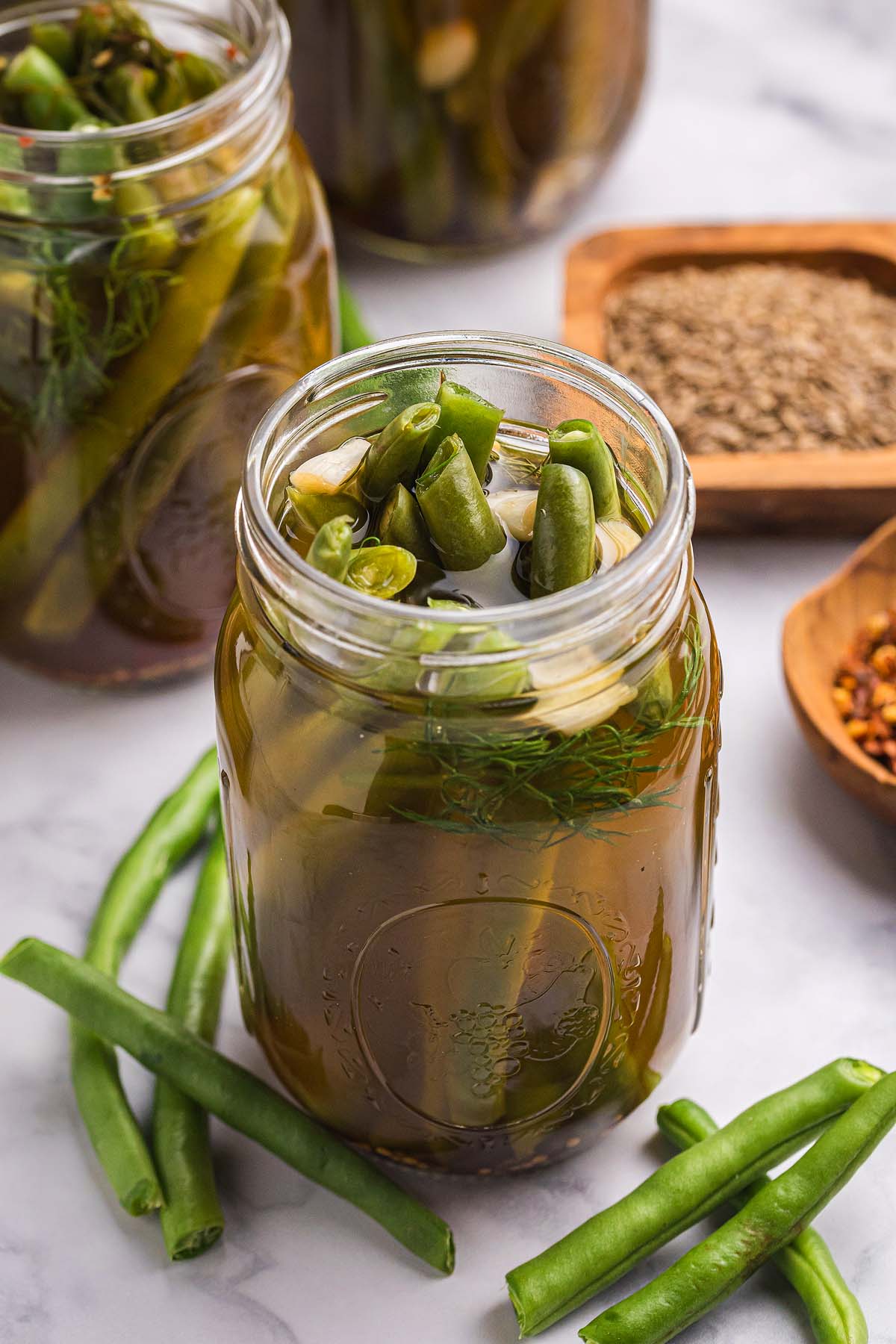 Jar of pickled green beans, garlic, and dill on a marble surface with fresh pickled green beans and spices nearby.