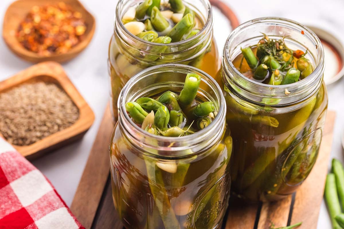 Three jars filled with pickled green beans and garlic sit on a wooden surface, surrounded by bowls of fragrant spices.