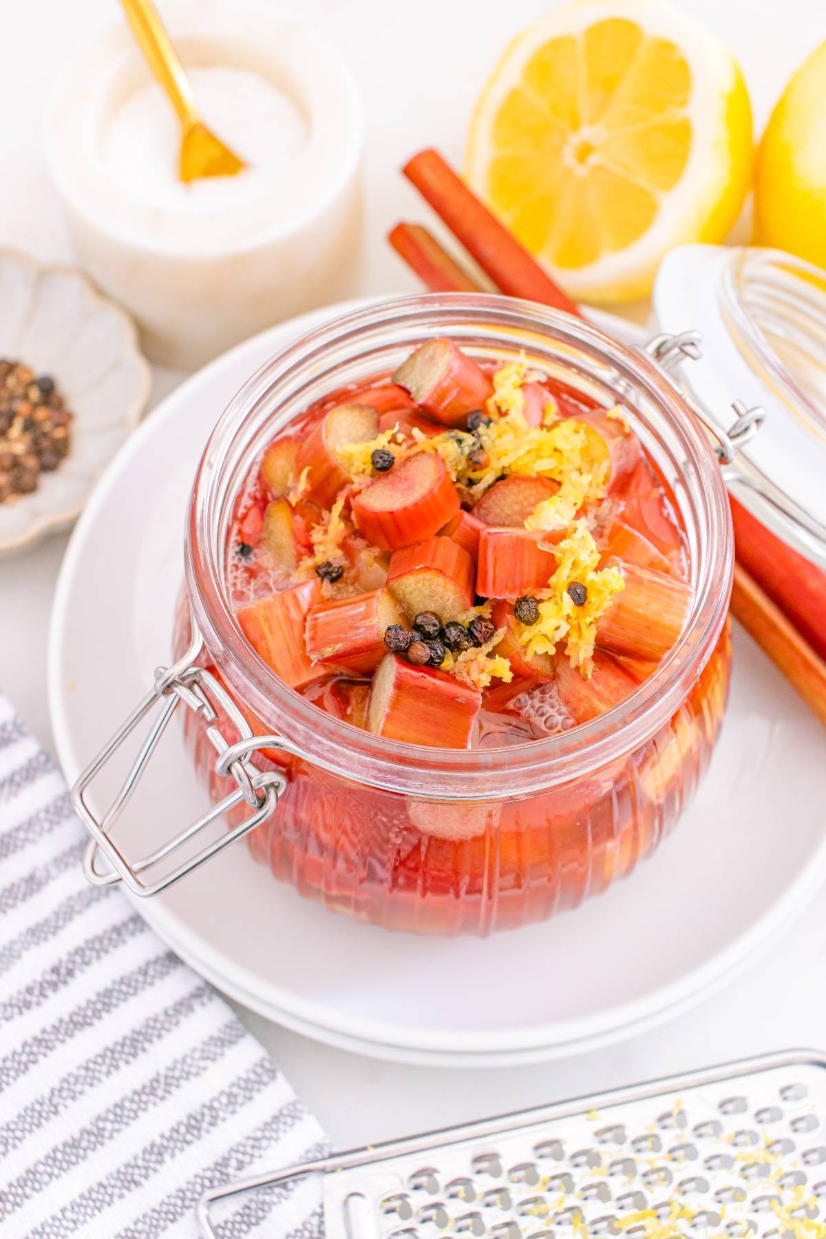 A jar of pickled rhubarb with spices and lemon zest, surrounded by lemons, cinnamon, and a striped towel.