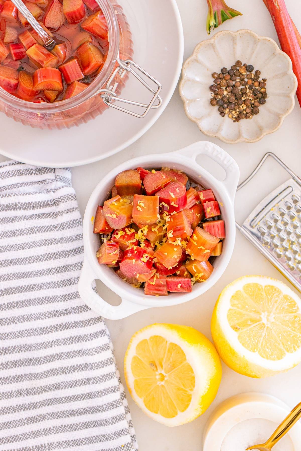 Small bowl of chopped rhubarb with lemon zest, surrounded by lemons, peppercorns, and a striped towel.