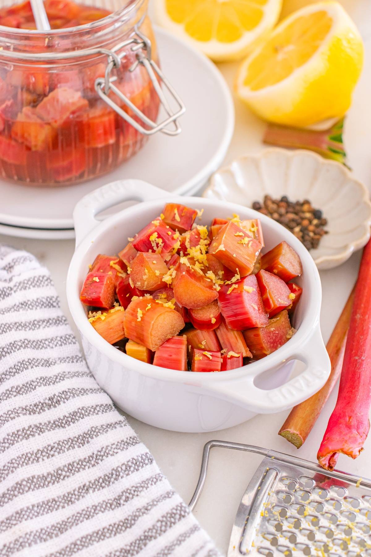 A white bowl of diced rhubarb topped with lemon zest, surrounded by lemon, spices, and a striped towel.