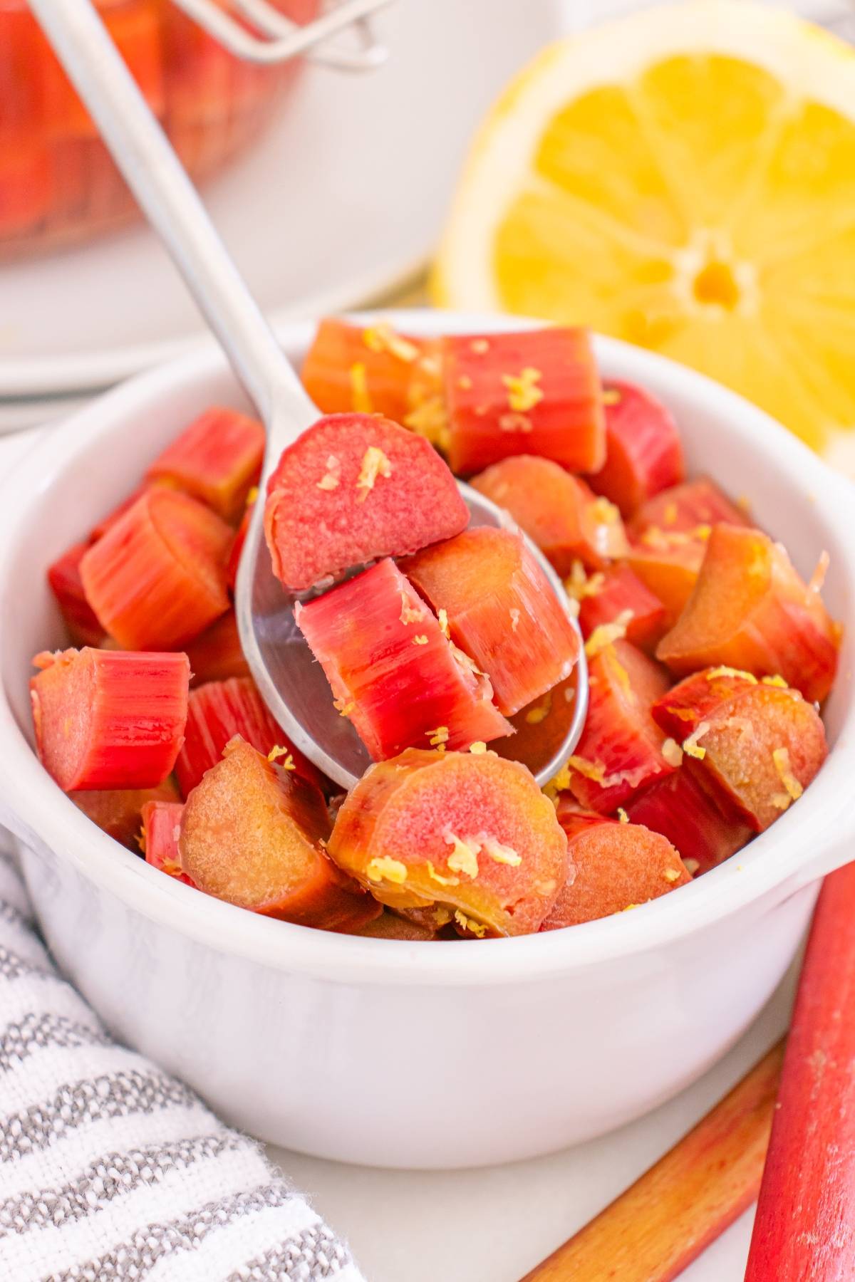 A spoon scoops chopped rhubarb pieces with lemon zest from a white bowl, with a lemon in the background.