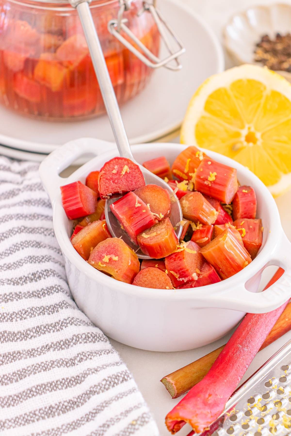 A white bowl of chopped rhubarb with lemon zest, a spoon, and sliced lemon beside it.