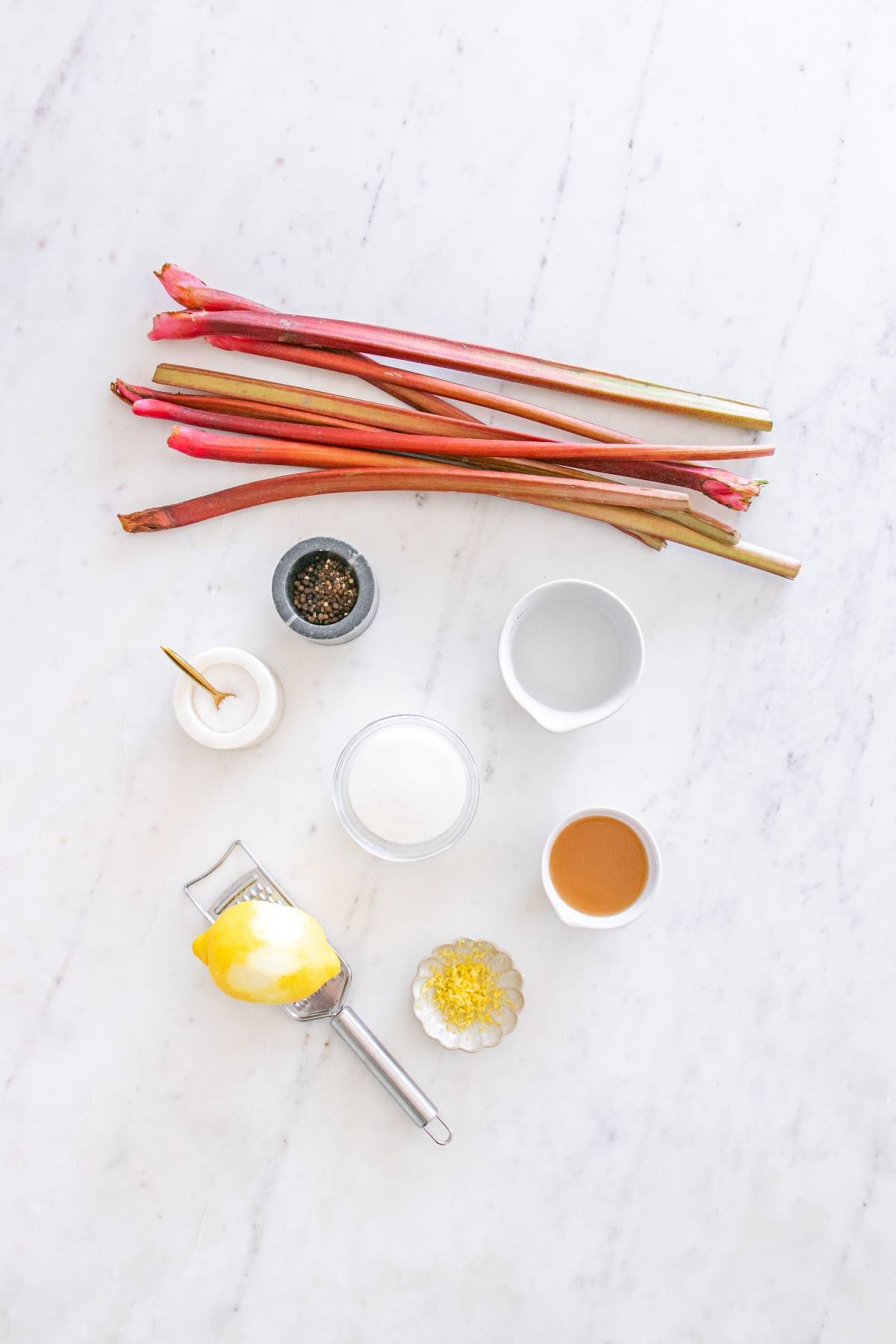 Rhubarb stalks, lemon, spices, sugar, and liquids arranged neatly on a white marble surface.