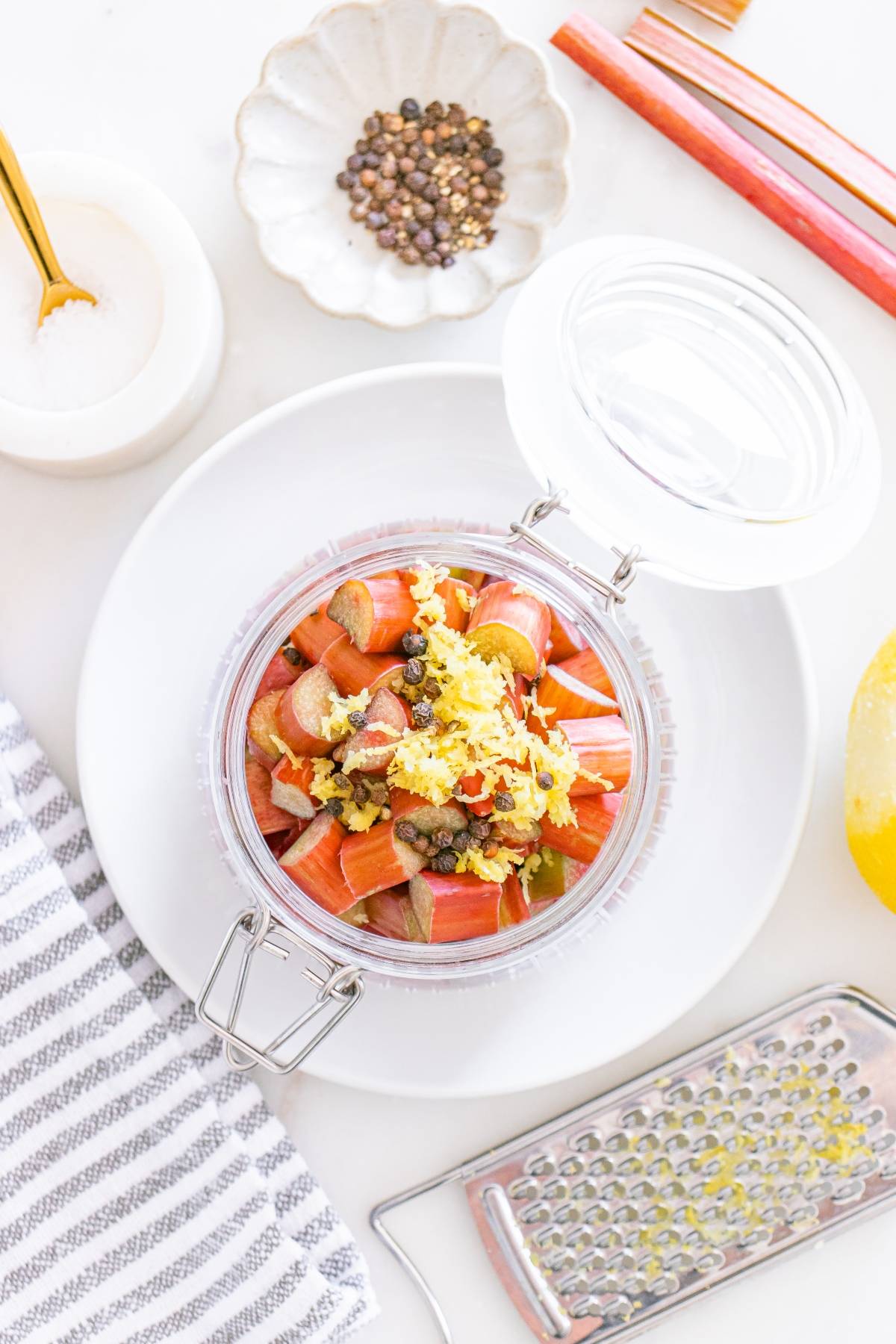 Jar of chopped rhubarb, grated ginger, and spices on a white plate with pepper, salt, and a grater nearby.