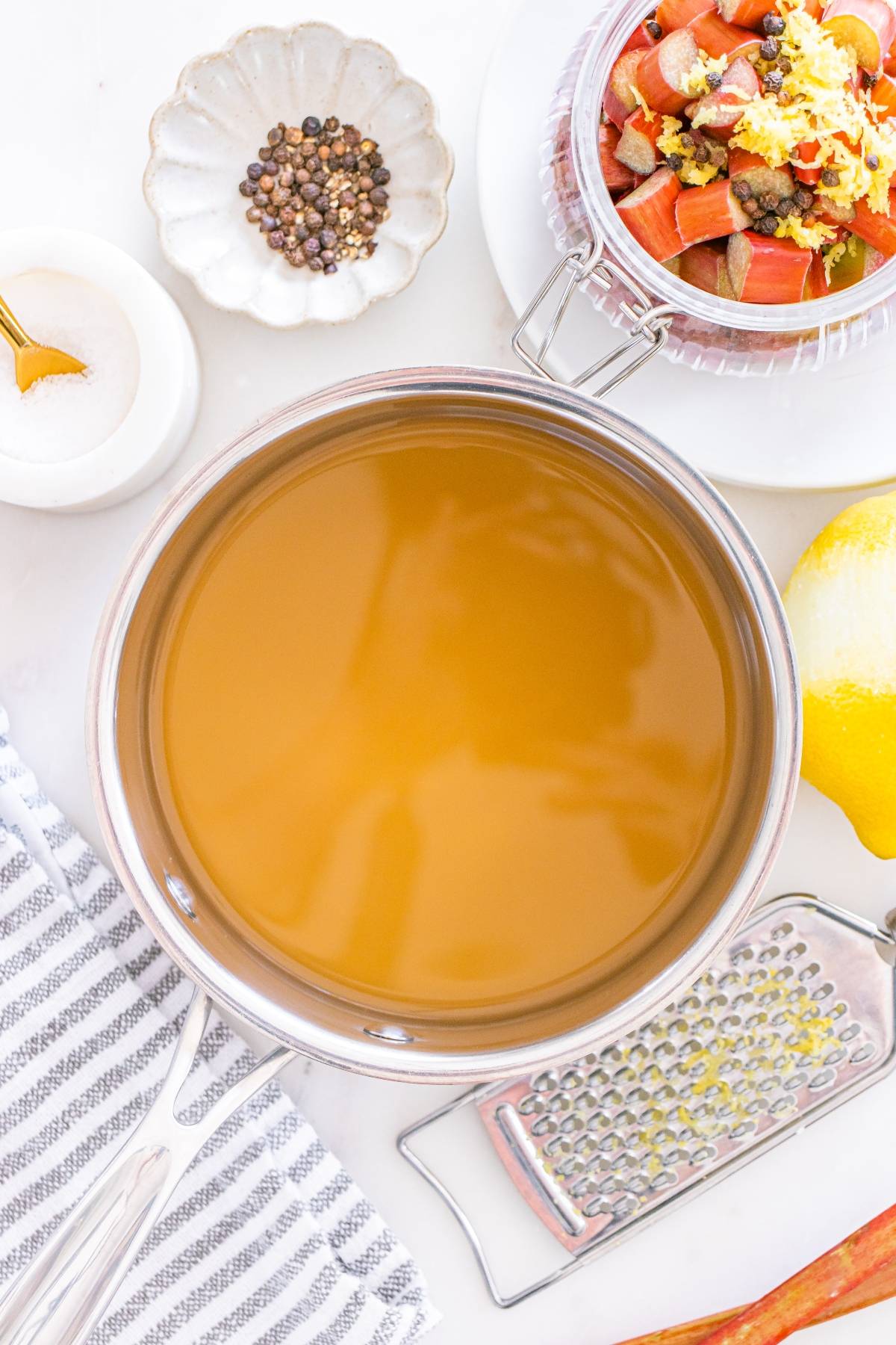 A pot of golden liquid surrounded by peppercorns, salt, rhubarb, a lemon, and a grater on a white surface.